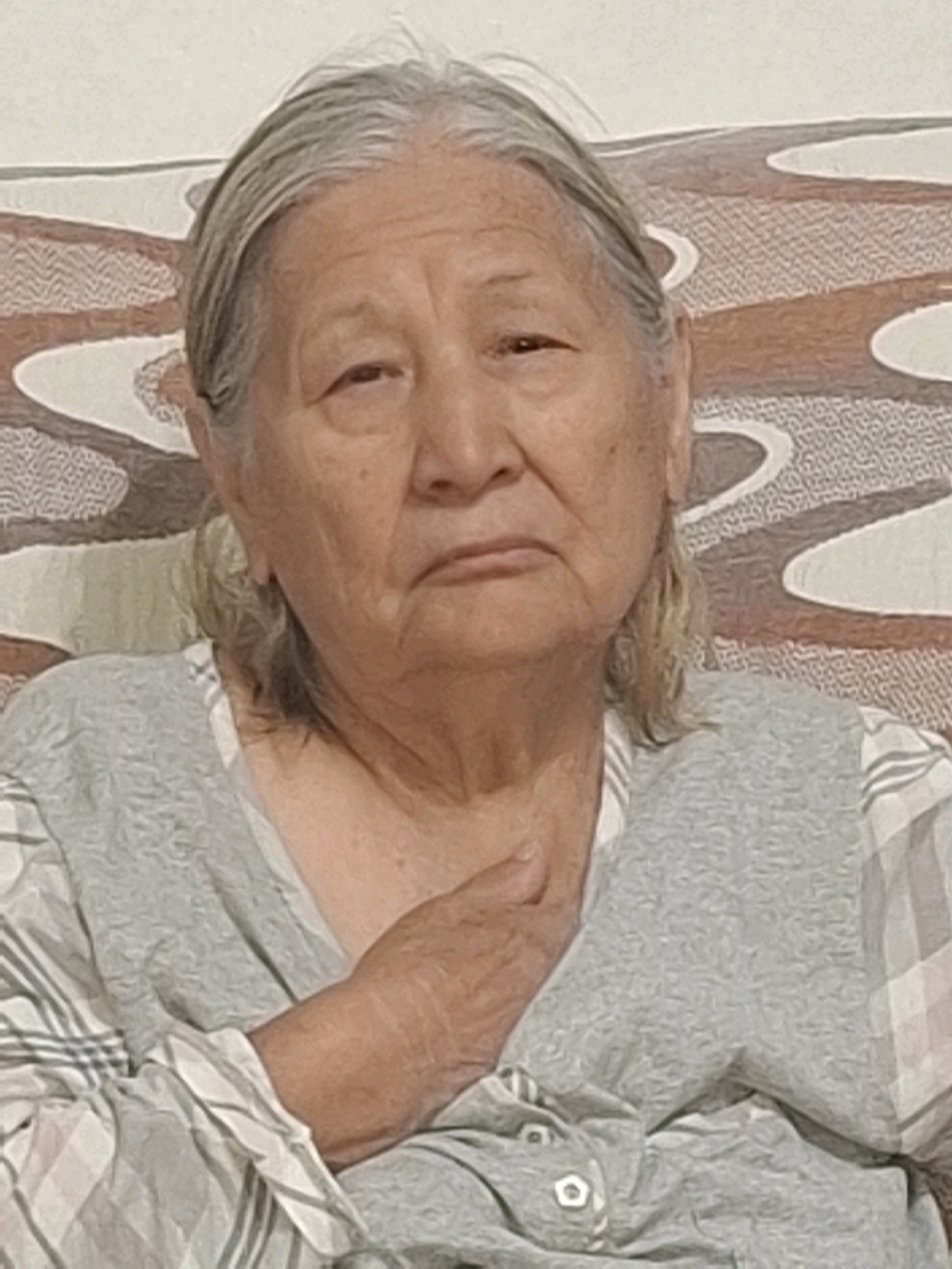 A headshot of an elderly woman with gray hair pulled back, a neutral expression, and her right hand resting on her chest. She is wearing a gray vest over a plaid shirt. The background appears to be a patterned sofa. The image was taken indoors in Almaty, Kazakhstan.