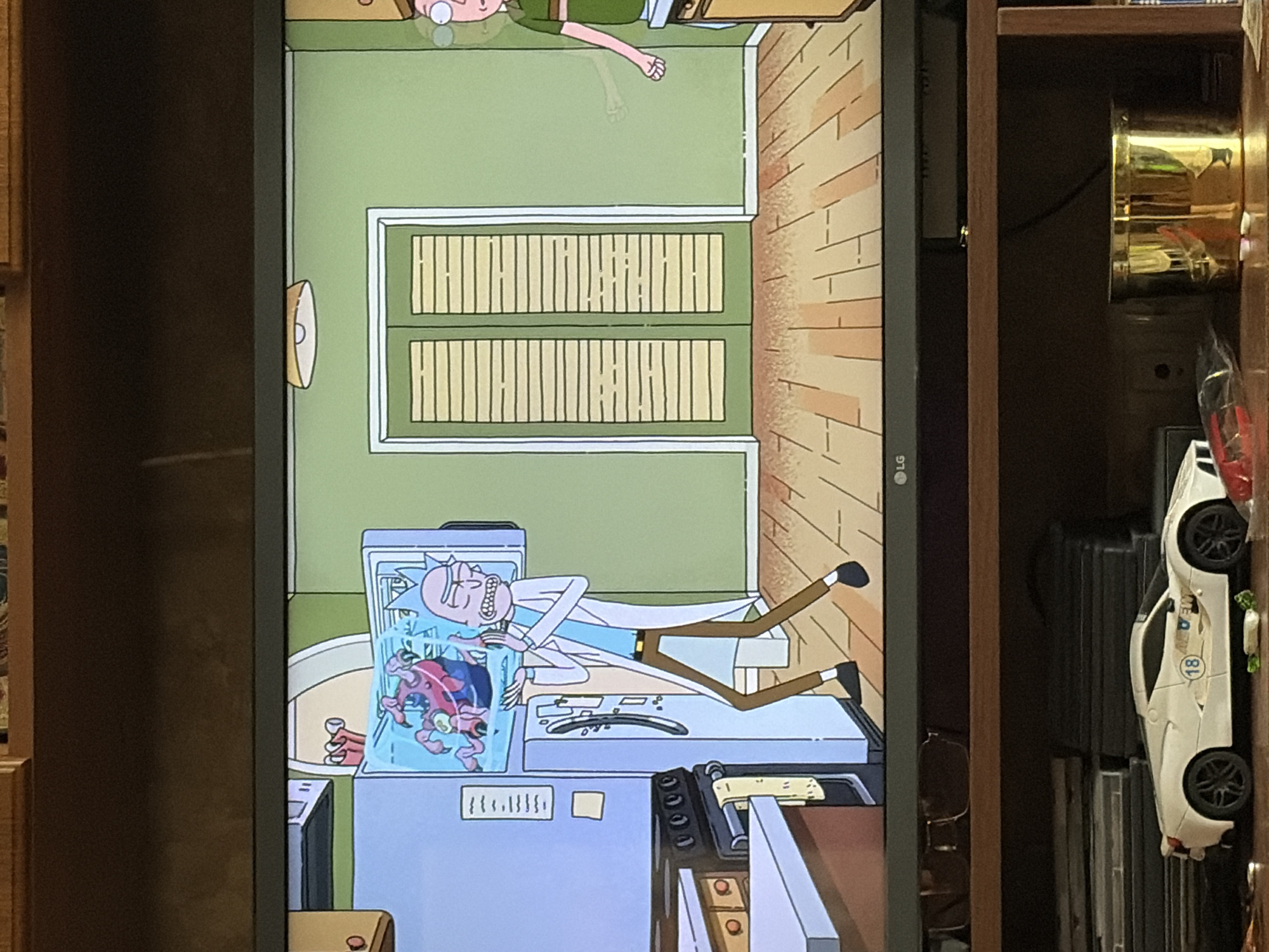 A television screen displays a scene from the animated show Rick and Morty. In the foreground, Rick Sanchez, a bald, mad scientist with blue skin and messy hair, stands in a kitchen wearing a lab coat. He is gesturing with his hands towards a refrigerator, which has a transparent compartment showing a large, red, multi-limbed alien creature. In the background, double doors with horizontal blinds are visible, as well as a faint, translucent figure of Morty Smith. The television is placed on a wooden shelf unit below a wooden cabinet. On the shelf below the television are various items, including a white toy sports car with the number 18 on its side, a stack of DVDs, and a golden container. The lighting in the room is dim. The scene appears to be indoors, likely a residential setting.