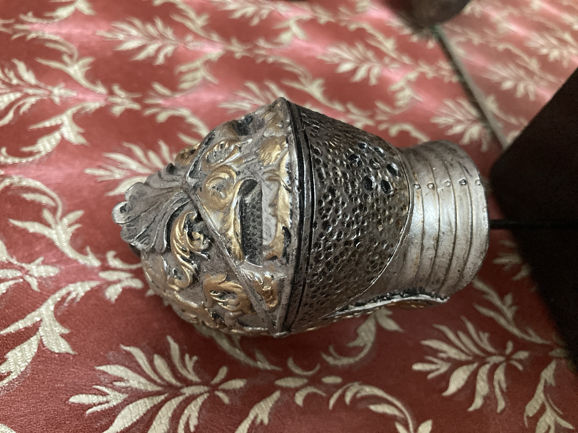 The image shows a close-up of an ornate, metallic object, possibly a decorative finial or a piece of costume jewelry, resting on a textured red fabric with a cream-colored floral pattern. The object is predominantly silver-toned with gold accents. One side of the object features intricate raised detailing with a swirling, leaf-like motif, highlighted in gold. The other side is textured with a repeating pattern of small, raised dots, resembling hammered metal or chainmail. This section also shows horizontal banding with circular indentations. A vertical seam or division is visible between these two distinct decorative styles. The object appears to be solid and substantial. The background fabric is a rich red, with the cream pattern appearing as stylized branches and leaves, creating a slightly raised, brocade-like effect. The lighting suggests an indoor setting, with soft illumination. There is no visible text in the image. The overall impression is one of detailed craftsmanship and possibly historical or fantasy-inspired design. The object is the sole focus, with no people or discernable environment beyond the fabric surface.