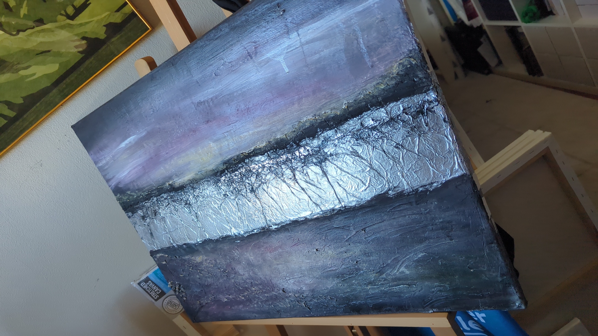 The image shows an abstract painting with textured elements, likely oil or acrylic on canvas, resting on an easel. The painting features a horizontal composition with a band of crinkled, metallic silver material across the middle, contrasting with darker, textured areas above and below. The upper section has washes of purple, grey, and hints of yellow, suggesting a sky or atmospheric effect. The lower section is a deep, textured grey with subtle purple undertones. The painting is displayed in what appears to be an indoor setting, possibly an art studio or a room within a residence. In the background, shelves filled with art supplies, canvases, and other objects are visible, along with a wooden floor. Another framed painting with abstract green shapes is partially visible on the left. The overall impression is one of artistic creation and display. The context of Kløfta, Norway, is not visually discernible from the image itself.