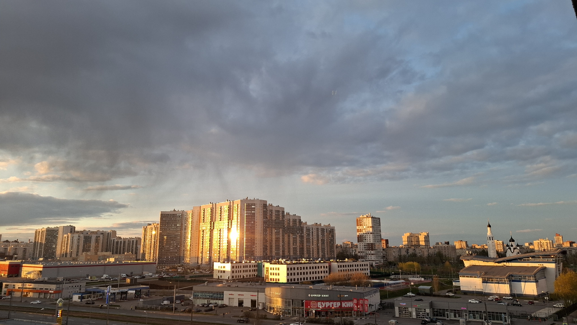The image captures an expansive urban landscape in Saint Petersburg, Russia, at golden hour or late afternoon. The sky presents a dramatic contrast: heavy, dark gray clouds dominate the upper left, suggesting rain or stormy weather, with faint streaks possibly indicating distant precipitation. To the right, the clouds lighten, revealing patches of blue sky and a warm, golden sunlight breaking through.
This sunlight strikingly illuminates the cityscape below. Several large, modern residential buildings in the midground and background glow with an intense golden reflection on their facades and windows, standing out against the darker sky. Below these towering apartments, the scene features various commercial establishments. A gas station is visible, along with low-rise buildings housing retail stores and what appears to be a car service center.
Prominent architectural features include a distinctive white and blue building, likely a public facility, and further to the right, a traditional Russian Orthodox church with white walls, a black onion dome, and a slender spire, characteristic of the region. Roads with scattered vehicles and parking lots indicate an active urban environment. Sparse trees, some showing early autumn colors, are also present.
Visible text includes a clear "БУРГЕР КИНГ" (Burger King) sign on a commercial building. Other Cyrillic signs present are "КУЗОВНОЙ РЕМОНТ" (Body Repair) and "ШИНОМОНТАЖ" (Tire Fitting) on a building in the lower left, and "СПОРТИВНЫЙ ЗАЛ" (Sports Hall) on the white and blue building. The overall mood is one of dynamic natural light over a busy city.