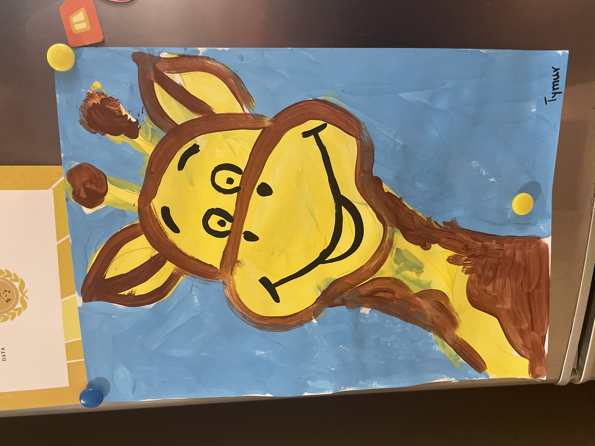 A close-up shot shows a child's painting of a giraffe's head and neck on a bright blue background. The giraffe is depicted with a large yellow face, brown ears, and brown spots on its neck. It has a wide, smiling mouth and simple black eyes and nostrils. The painting is attached to a metallic surface, likely a refrigerator, with two blue magnets and one yellow magnet. There is also a small, colorful house-shaped decoration and a fragment of a certificate or document visible at the top. The artist's signature, "Tymur," is visible in black marker in the lower right corner of the painting.