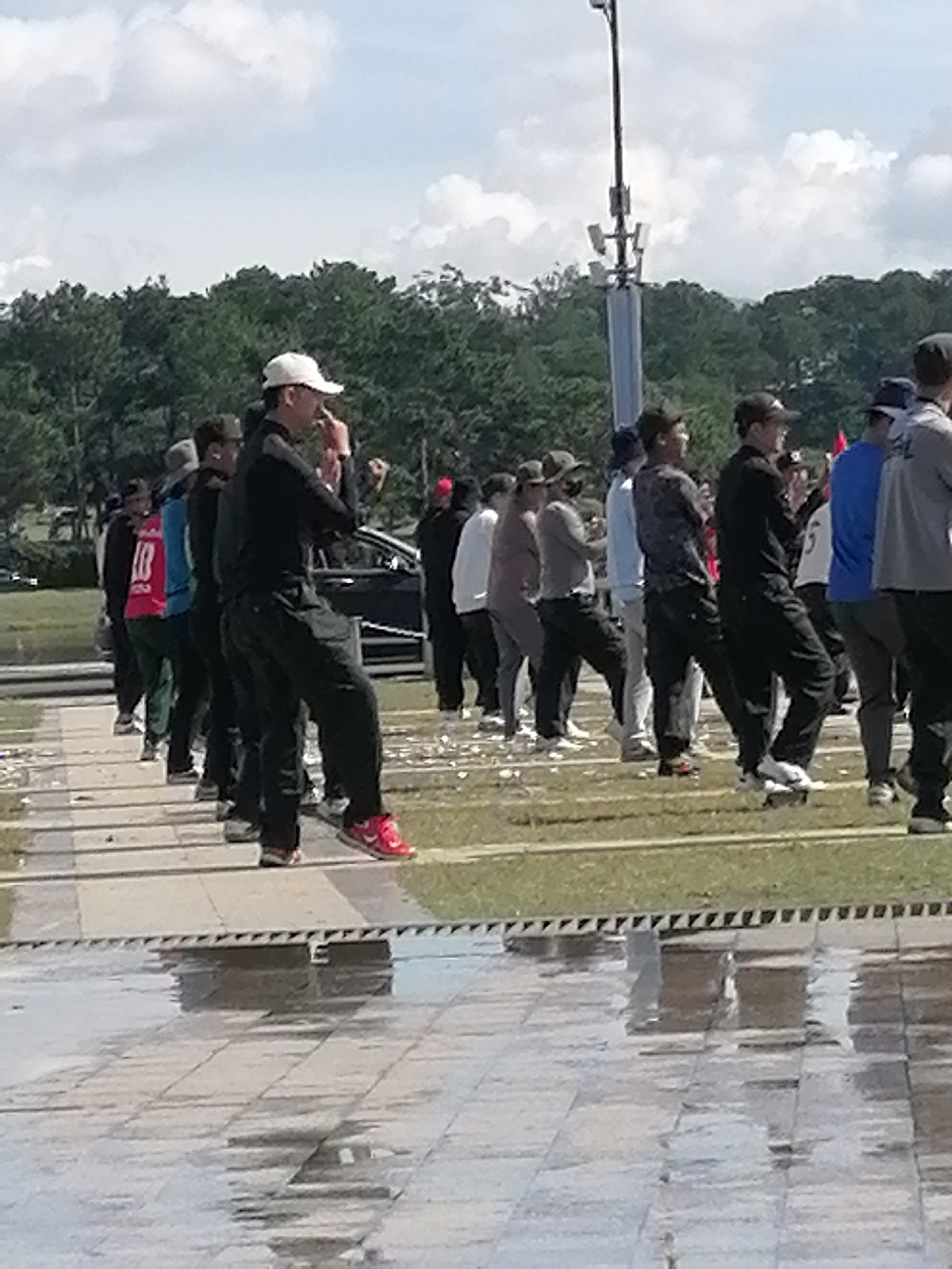 A group of approximately 15 men, dressed in casual athletic wear such as tracksuits and baseball caps, are engaged in what appears to be an outdoor exercise or activity on a paved area. The men are in various stages of movement, with some standing still and others in mid-stride or with arms raised. The ground is wet and reflects the surroundings, suggesting recent rain. In the background, there is a line of trees, and beyond that, a cloudy sky. A tall, multi-tiered pole with lights or antennas is visible on the right side of the frame. A dark-colored vehicle is parked in the middle ground, behind some of the participants. The overall scene suggests an organized gathering in an open outdoor space. The presence of lush trees and the cloudy sky are consistent with the natural environment of Da Lat, Vietnam, known for its cool climate and scenic landscapes.