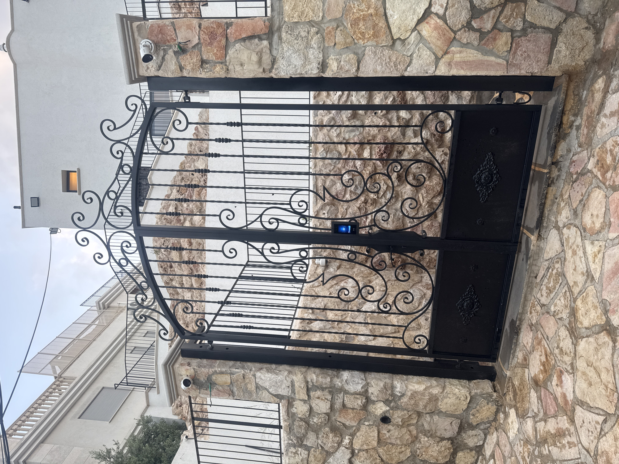 The image shows a black wrought iron gate in Ramat Gan, Israel. The gate is set between two stone pillars. The gate has an ornate design with swirling patterns and vertical bars. A small, illuminated blue device is attached to the gate. In the background, a multi-story building with a white facade is visible. A surveillance camera is mounted on the stone pillar above the gate. A second surveillance camera is mounted on the building facade. The gate is set on a paved surface. The sky is visible in the upper left corner of the image.