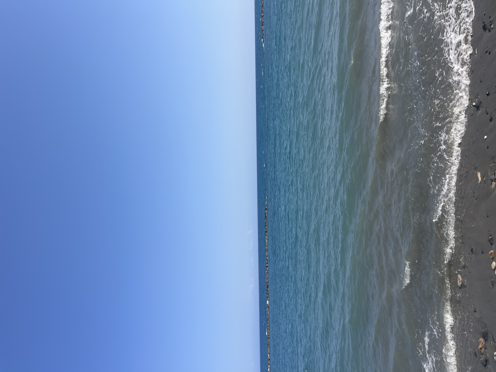 A wide, horizontal, eye-level shot captures a serene view of the Mediterranean Sea under a clear blue sky. The foreground features a dark, pebble-strewn beach, with gentle waves lapping at the shore, creating white foam. In the middle ground, the calm, blue-green water stretches towards the horizon, interrupted by a man-made stone breakwater that extends horizontally across the scene, with small white caps visible near it. The background is dominated by the vast expanse of the clear, bright blue sky. The overall impression is one of tranquility and natural beauty.
