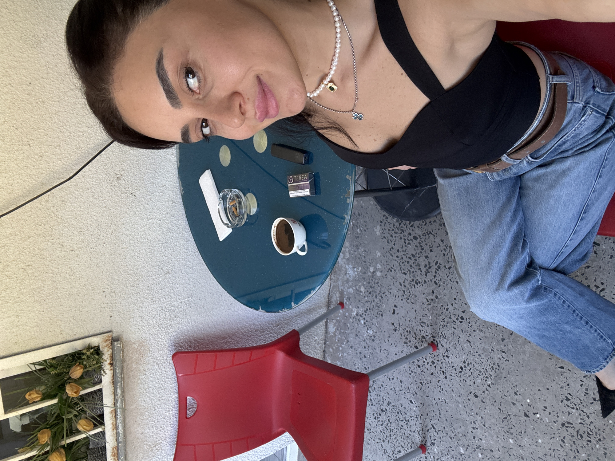 A woman sits at a small, round, blue table on a concrete patio. She is wearing a black tank top and blue jeans. She has dark hair and is looking at the camera. On the table, there is a cup of coffee, an ashtray with cigarette butts, a pack of cigarettes, and a vape device. To her left, there is a red chair. In the background, there is a white textured wall with a window box containing yellow flowers. The scene is outdoors and appears to be in Tbilisi, Georgia.