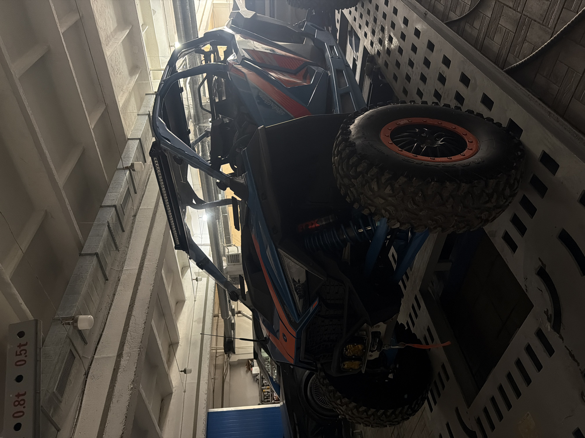 The image shows an all-terrain vehicle (ATV) parked in a garage or storage facility. The ATV is blue and orange, with large, knobby tires. It is parked near a wall with a regular pattern of square indentations. The location appears to be an indoor space, with concrete walls and a high ceiling, likely a commercial or industrial building. There is a sign on the wall indicating weight limits, suggesting this might be a storage or maintenance area. The city and country are not discernible from the image.