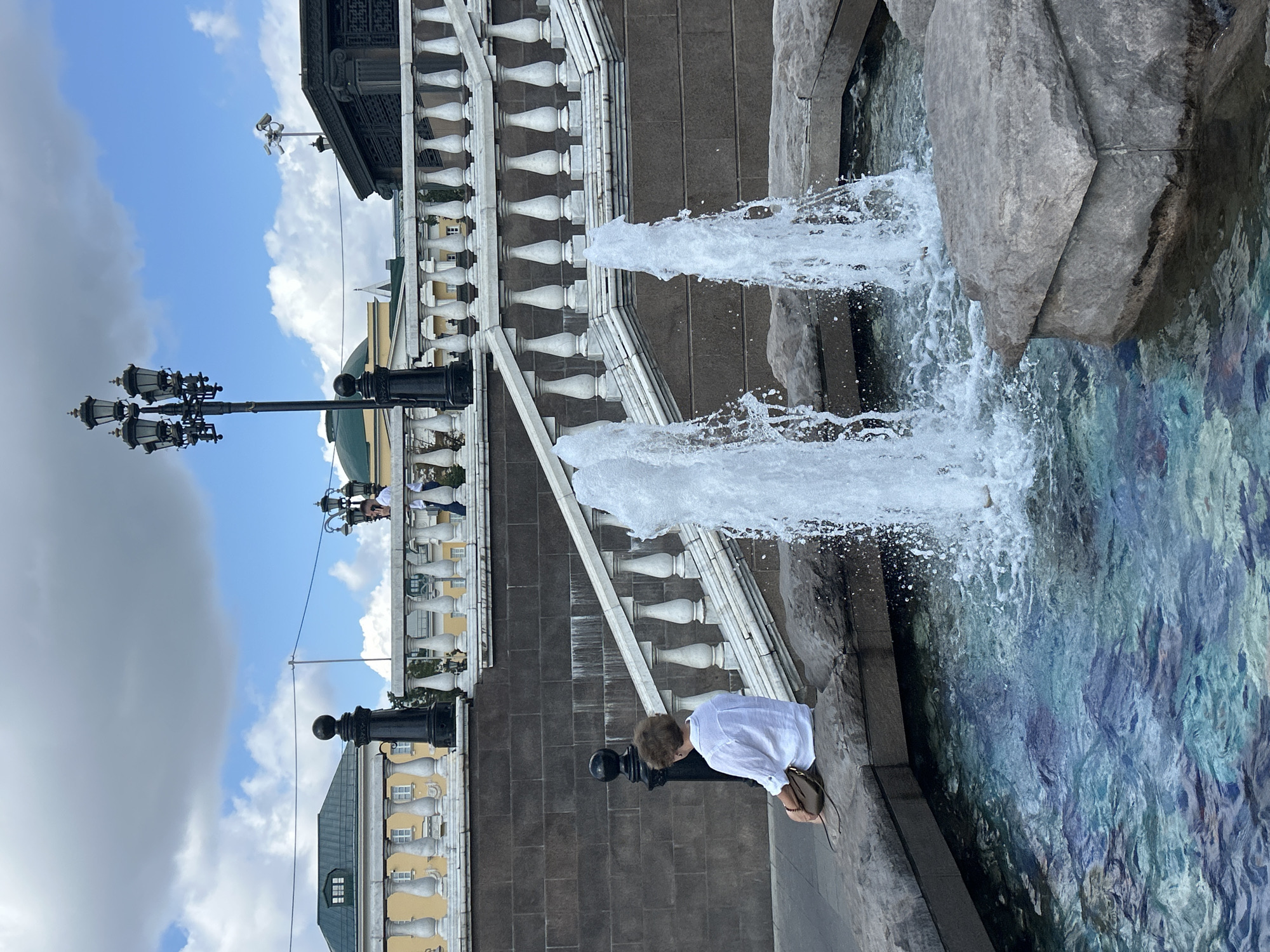 A lone person wearing a white shirt sits on the edge of a stone fountain in an urban outdoor setting. The person has curly brown hair and is facing away from the camera, with a small brown bag resting beside them. Two fountains are active in the foreground, spraying water upwards into the air. The fountain has a stone basin with large, grey rocks. Behind the fountain, a wide stone staircase with white balustrades leads up to a raised level. A dark stone wall forms the base of the staircase. In the background, there are several buildings with yellow facades and green roofs, along with a black lamppost. The sky above is partly cloudy with blue sky visible. The location is an unknown city.