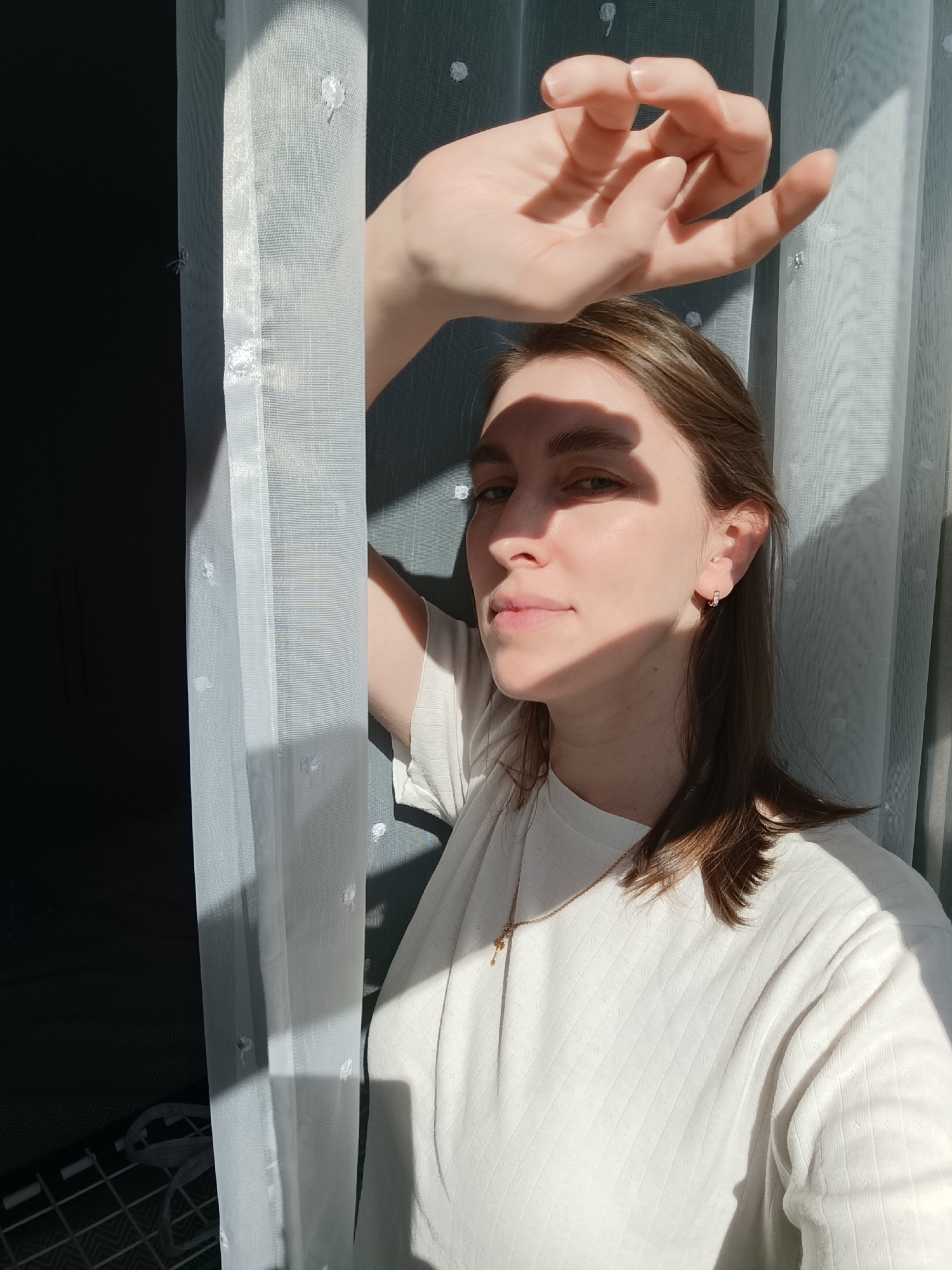 The image displays a close-up indoor portrait of a female individual, located in Chaikovsky, Russia. The subject, with fair skin and light brown hair, is positioned with her upper torso and head visible. She wears a light-colored, ribbed long-sleeved top and a thin gold-colored necklace with a small pendant. A small gold-colored hoop earring is visible in her right ear. Her right hand is raised above her head, partially obscuring her forehead and casting a distinct shadow across the upper portion of her face. Her eyes are light-colored, and she maintains direct eye contact with the viewer. Strong sunlight enters from the right, creating bright highlights and sharp shadows, including a vertical shadow line from a curtain on the left side of the scene. The background consists of a sheer white curtain with small textured dots, which is illuminated by the sunlight and partially filters the light. Behind the curtain, a darker, possibly patterned surface is visible.