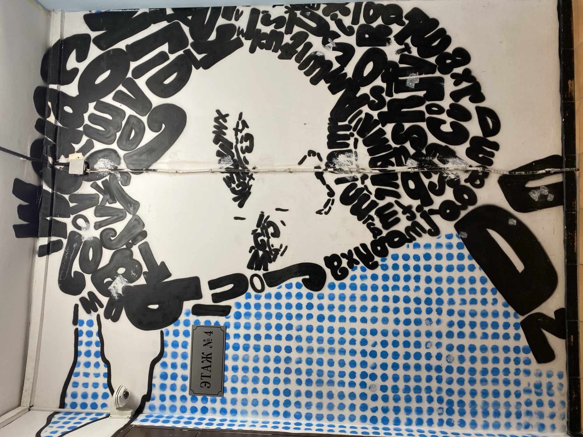 The image captures a vibrant mural painted on a white wall, likely within an indoor setting such as a hallway or stairwell in Moscow, Russia. The central subject is a stylized depiction of a person's head and shoulders, shown in profile facing right.
The most prominent feature is that the hair and the outline of the head are formed by a dense arrangement of numerous black, bubble-like letters and numbers of varying sizes and orientations. Visible words and phrases within this textual "hair" include "COVID," "19," "MASK," "STAY HOME," "DISTANCE," "NOAM," "VIRUS," "BREAK," "SAFE," and "HEALTH," strongly suggesting a theme related to the COVID-19 pandemic and public health messages. The face itself is rendered with minimalist black lines.
Below the head, the person's upper body or clothing is represented by a striking pattern of bright blue polka dots against a white background, outlined in black. Several real-world elements are integrated into the mural: electrical wires run horizontally across the mid-section of the image, partially obscuring the artwork, and a small white junction box is attached to the wall. A small white security camera is also visible near the bottom left. A grey rectangular plaque with the Russian text "ЭТАЖ №4" (ETAZH No. 4, meaning Floor No. 4) is affixed to the wall within the blue polka-dot section. The mural appears to be a piece of thought-provoking street art or social commentary.