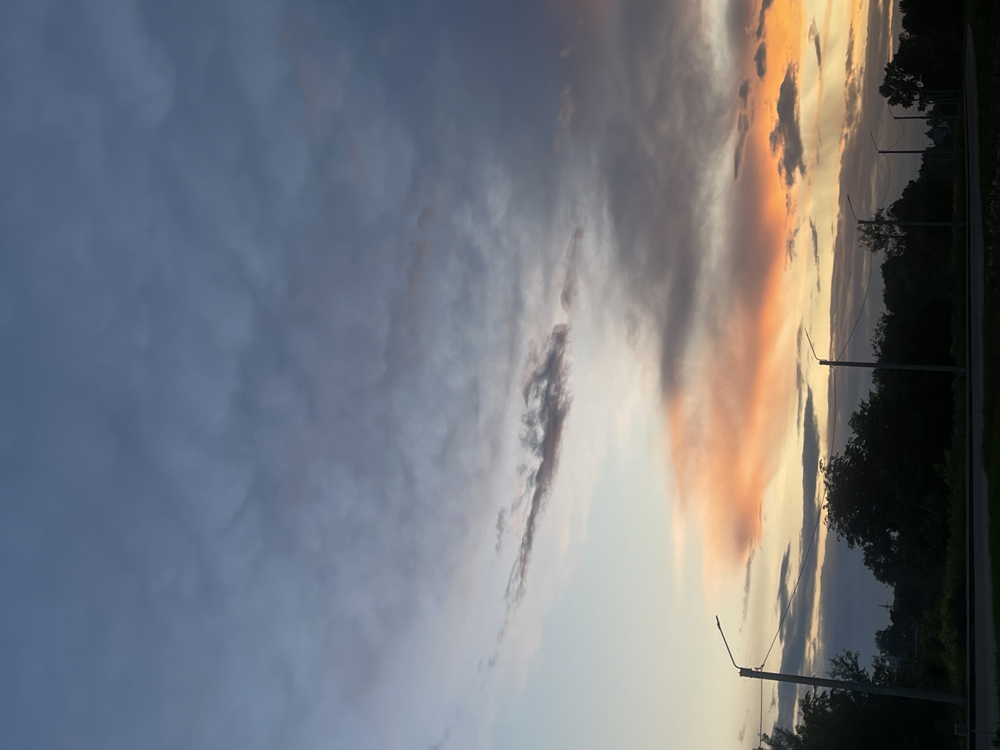 This image, taken in Votkinsk, Russia, captures a striking sunset or sunrise over a landscape featuring natural and man-made elements. The sky dominates the scene, displaying a dramatic contrast of colors. On the right side of the frame, clouds are brilliantly illuminated by the sun, showcasing warm hues of orange, pink, and yellow. Interspersed within these bright clouds are darker, more defined cloud formations. Towards the left, the sky transitions into cooler tones of blue and gray, with lighter, wispy clouds creating a layered effect.
Along the bottom of the image, a dark silhouette of trees and foliage forms a natural boundary, suggesting a ground level. Several street lights, appearing as slender, dark poles with horizontal light fixtures, are visible among the trees, indicating a developed area, possibly alongside a road or path. A dark, narrow strip, likely a road or pavement, runs along the very bottom edge of the frame. The time of day is clearly either dawn or dusk, characterized by the intense, colorful illumination of the clouds. The weather appears partly cloudy. No visible text is present in the image.