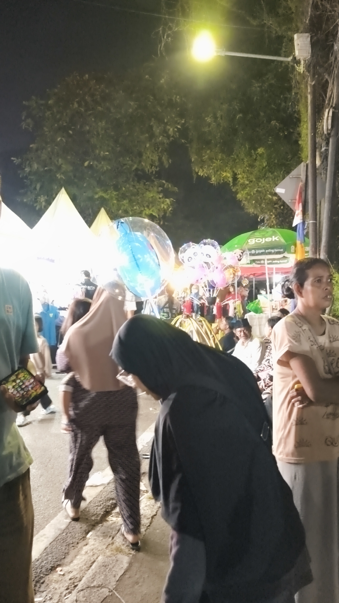 The image depicts a nighttime scene at an outdoor market or fair in South Jakarta, Indonesia. In the foreground, several individuals are visible. To the left, a person wearing a light blue t-shirt and brown pants is holding a tablet. In the center, a person in a brown hijab and patterned pants is walking away from the camera. To the right, a person wearing a black hijab is leaning forward. Further to the right, a woman in a light brown t-shirt stands with her arms crossed, looking to the right.
The background is populated with various stalls and tents, some of which are illuminated by bright lights. A prominent green Gojek umbrella is visible, suggesting the presence of ride-hailing services or related vendors. Large, colorful balloons, including a blue one and pink ones with panda faces, are being sold or displayed. Trees with dense foliage are present, partially illuminated by a street lamp hanging from a pole on the right side of the frame. The ground is paved and appears somewhat worn, with some debris present. The overall atmosphere suggests a lively, public gathering.