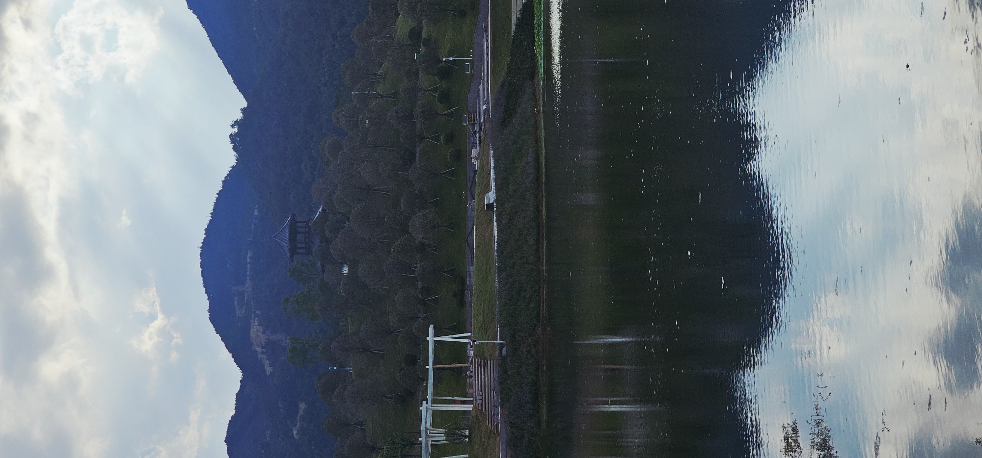 The image depicts a tranquil waterscape with a mountain backdrop, likely taken in the city of Chongqing High-tech Zone, China. In the foreground, a calm body of water reflects the sky and the surrounding landscape. The surface of the water is disturbed by ripples and dotted with small floating debris.
Beyond the water, a green embankment is visible, transitioning into a grassy hillside dotted with numerous trees. These trees appear to be deciduous and are in a state of dormancy, with bare branches. A modern white pavilion structure with a layered roof is situated on the left side of the embankment, providing a shaded area. Further up the hill, a traditional dark-colored pavilion with an ornate roof is nestled among the trees, partially obscured by their branches.
The middle ground is dominated by the densely planted trees that ascend the slope of the hill. The background consists of the dark blue silhouette of rolling mountains, covered in vegetation. The sky above is overcast, with patches of light breaking through the clouds, casting a soft illumination on the scene. The overall impression is one of a serene park or natural landscape, likely a public space designed for recreation and appreciation of nature. There are no discernible people in the image.