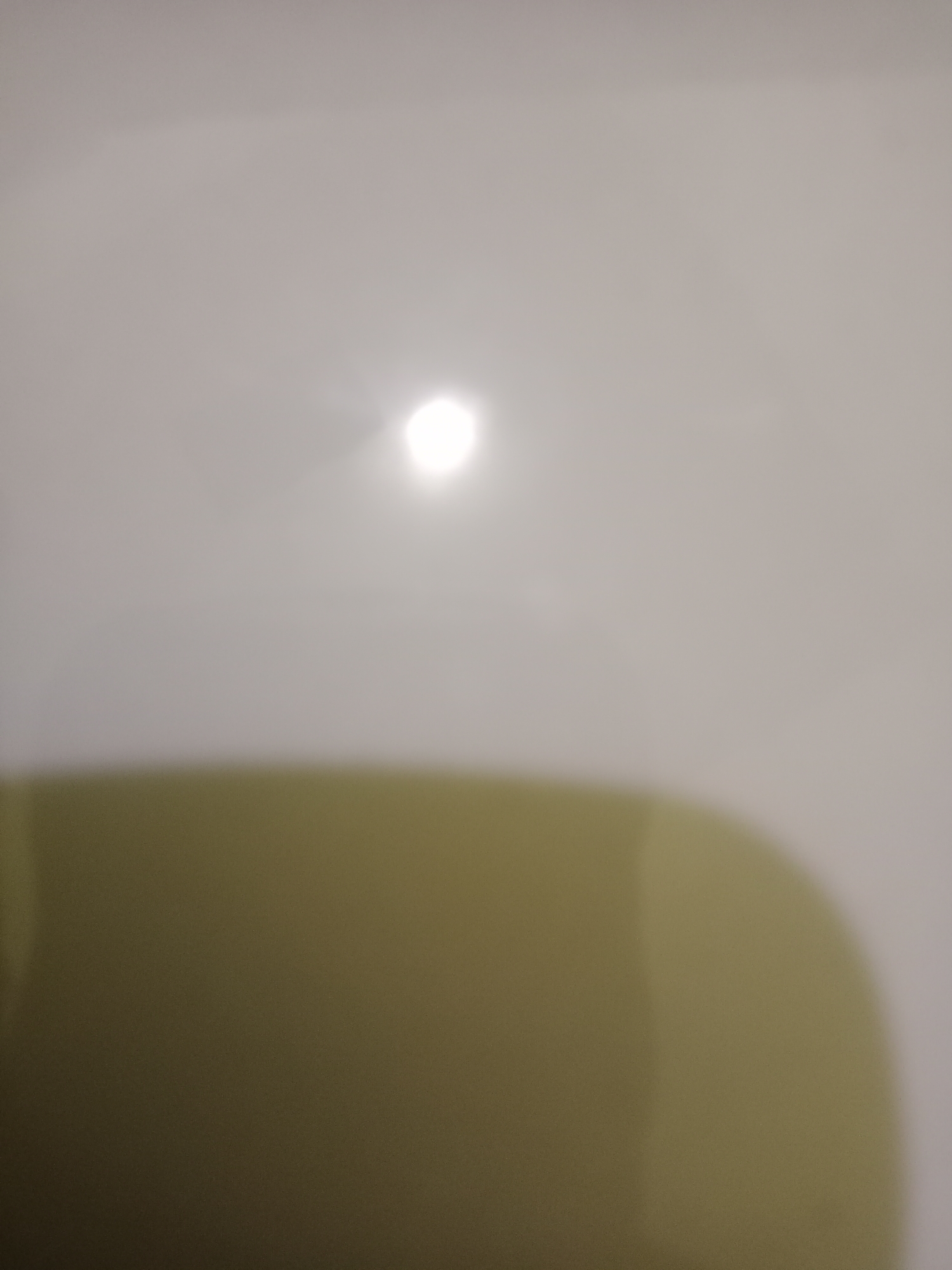 The image is a blurry, abstract depiction of a light source, possibly a camera flash, illuminating a plain, off-white surface. Below this, a large, blurred area of olive green occupies the lower half of the frame. The lighting suggests an indoor environment, but no specific location or objects are identifiable beyond the illuminated surface and the colored area. There are no people or any identifiable features that could place the image in West Jakarta, Indonesia.