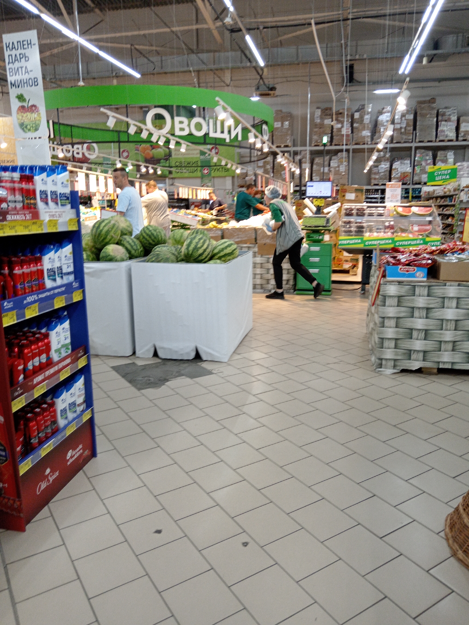 The image depicts the interior of a supermarket, likely in Belogorsk, Russia, given the context. In the foreground, a blue shelf unit displays various personal care products, including red and white bottles. To the right of this unit, a large white container is filled with watermelons. Several people are visible in the background. Two men, one in a light blue shirt and the other in a white shirt, appear to be working near the watermelons. A person wearing a green vest and dark pants is walking away from the camera towards a checkout area, carrying a white plastic bag. The environment is a typical supermarket aisle with tiled flooring, bright overhead lighting, and shelving units filled with merchandise. Signs in Russian are present, including one that reads "ОВОЩИ" (Vegetables). The overall scene suggests a busy retail environment.
