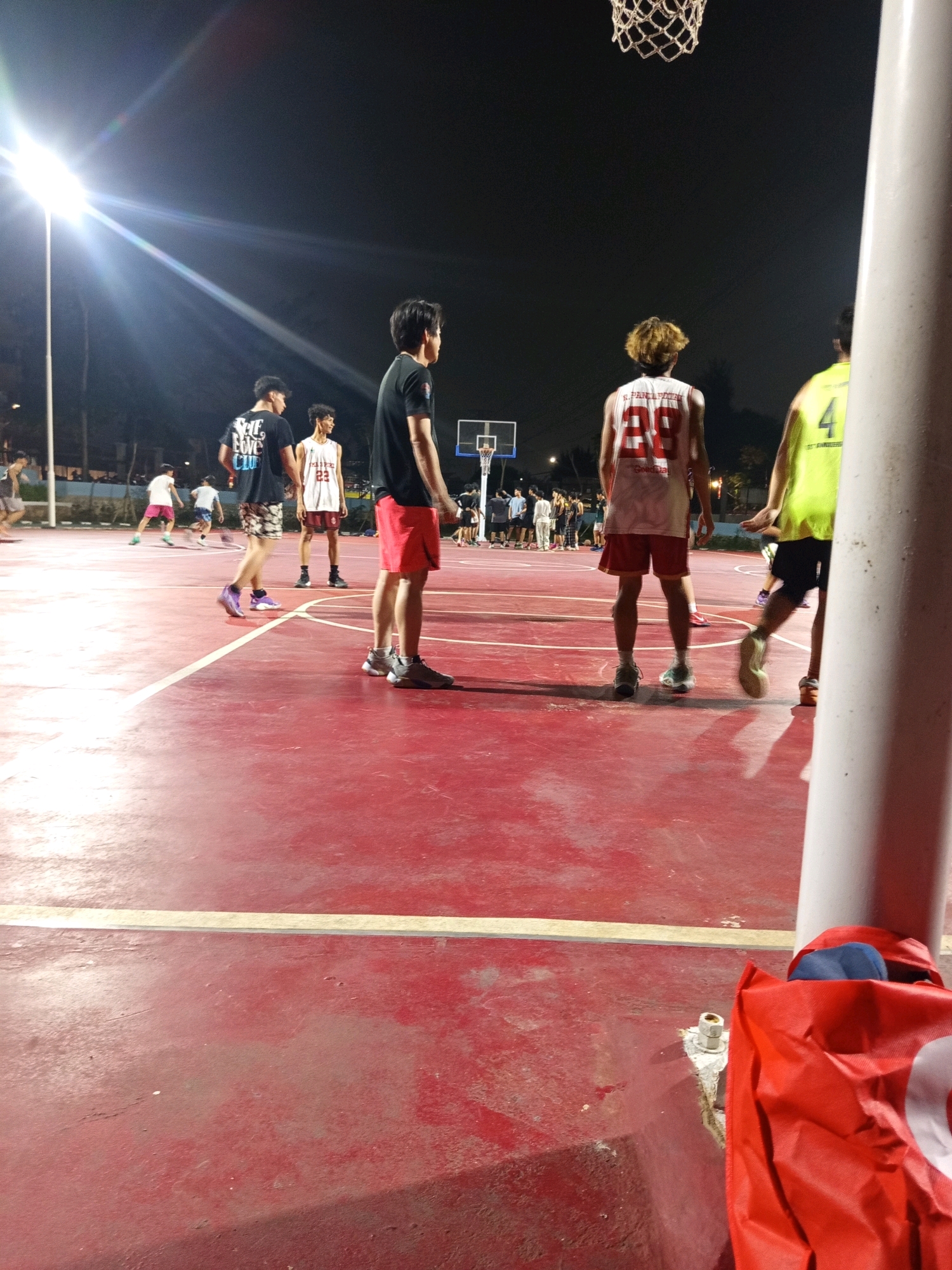 The image depicts an outdoor basketball court at night, illuminated by bright overhead lights, casting long shadows across the red court. Several individuals are visible, some actively playing or observing. In the foreground, to the right, a bright red bag and a white pole dominate the frame.
Further into the court, four young men are the most prominent subjects. Two are facing away from the camera, one in a white and red jersey with the number "23" and another in a bright yellow jersey with the number "4". Another man, wearing a black t-shirt and red shorts, is also facing away from the camera. To his left, a man in a black t-shirt and patterned shorts is turned towards the players, appearing to be observing the game.
In the mid-ground, other figures are visible engaged in basketball activities. To the left, a player in a white t-shirt and pink shorts is running, while other individuals are scattered around the court. In the background, a basketball hoop and backboard are visible, along with more people spectating near the court's edge. The overall atmosphere suggests a casual, night-time basketball game.
The scene is set in West Jakarta, Indonesia, indicated by the distinct style of the basketball court and the typical lighting found in public outdoor recreational spaces in the city.