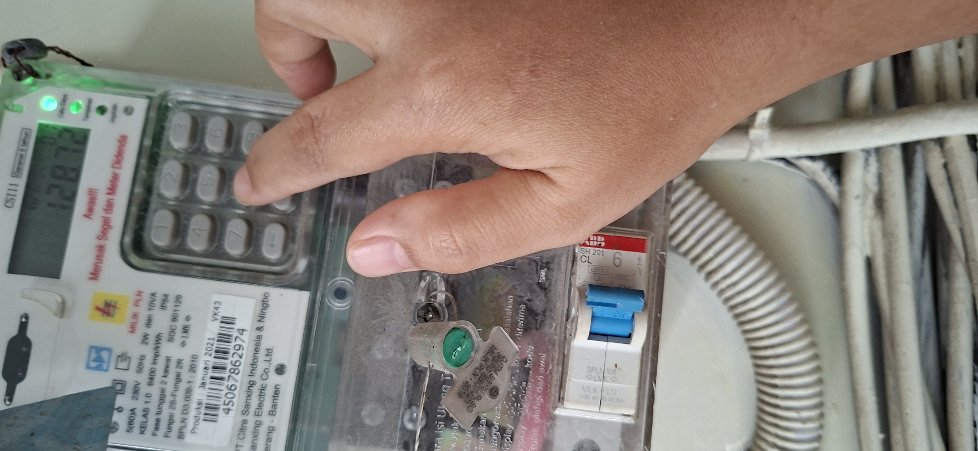 In this close-up shot, a person's hand is interacting with a digital electricity meter. The meter, identified as a CSI11 model, displays a reading of 128.72 kWh and shows a green light indicating power consumption. The meter has a keypad for inputting numbers, and the person's finger is pressing one of the buttons.
The meter is affixed to a wall, and visible on the meter's casing are various labels and specifications. One label indicates the manufacturer as "PT. Citra Sanxing Indonesia & Ningbo Sanxing Electric Co., Ltd.," with a production date of January 2021. Another label displays a serial number: "45067862974." Below the keypad, there is a circuit breaker with a blue toggle switch.
The environment appears to be indoors, with a white wall as the background. The lower portion of the image reveals bundled electrical wires and a corrugated conduit, suggesting the meter is part of a building's electrical system. The lighting is adequate, highlighting the details of the meter and the person's hand. The presence of a hand interacting with the meter implies the person is either checking the reading, inputting a token, or performing some other function related to the meter. The image does not provide specific information about the location being Surabaya, Indonesia, or any details about people other than the visible hand.