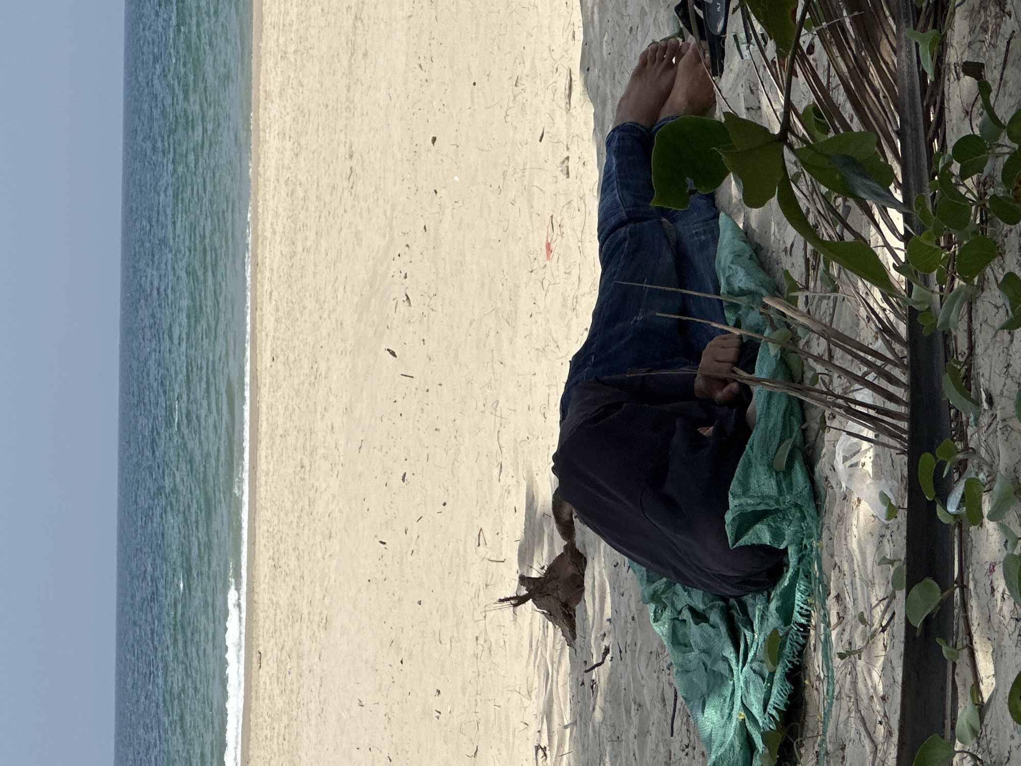 A person is sleeping on a green tarp on a sandy beach with the ocean in the background. The person is wearing blue jeans and a dark long-sleeved shirt, with their head tucked into their arms. The beach is sparsely littered with debris, including a piece of driftwood and some dried palm fronds. The ocean is calm, with a clear blue sky overhead. The scene appears to be located on My Khe Beach in Đà Nẵng, Vietnam.