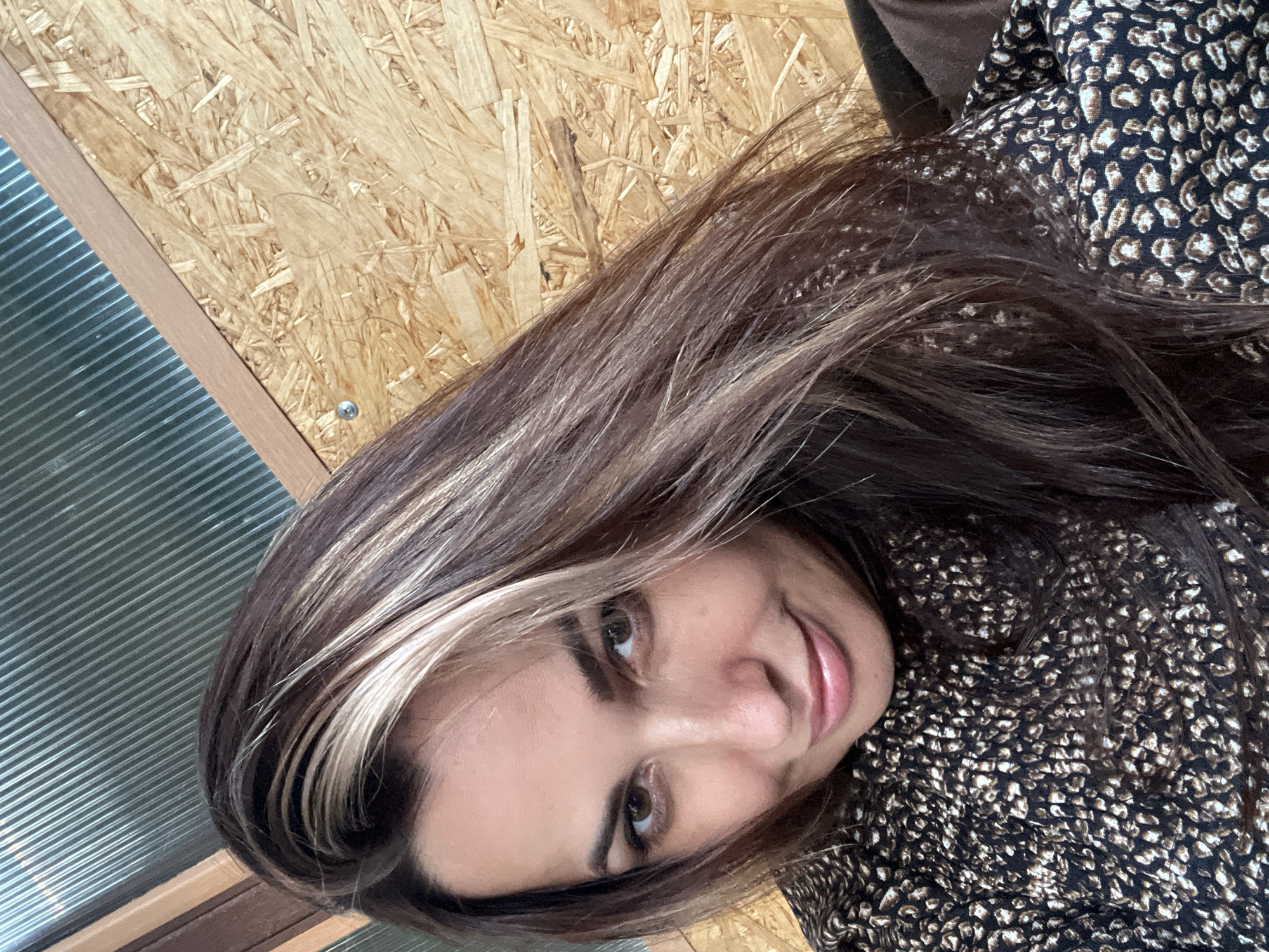 In a close-up, waist-up selfie, a woman with dark brown hair featuring blonde highlights looks directly at the camera with a slight smile. She is wearing a dark top with a light-colored leopard-like print. The background consists of a textured wooden wall and a section of corrugated metal sheeting, suggesting an indoor or possibly an outdoor shed or workshop setting. The lighting appears natural and somewhat diffused.