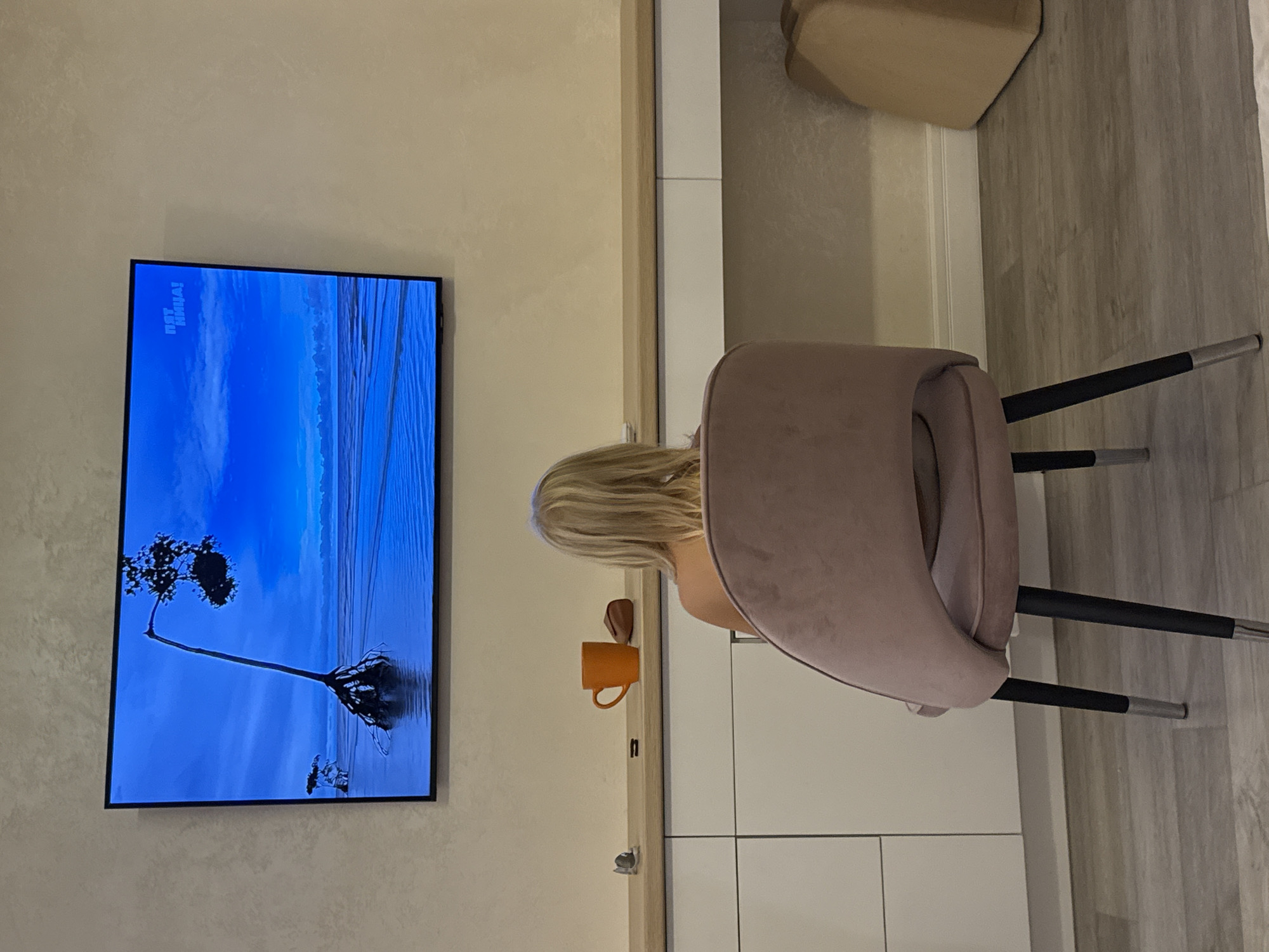 In a dimly lit room, a young child with blonde hair sits facing away from the camera, engrossed in watching a television screen. The television displays a nature scene with a stark blue sky and water, featuring a distinctive mangrove-like tree with exposed roots. A logo that reads "ПЯТНИЦА!" is visible in the upper right corner of the screen.
The child is seated in a pink velvet chair with black legs tipped with silver. The chair is positioned in front of a long, light-colored desk. On the desk, a bright orange mug, a small silver object, and a dark, amorphous object are visible. The desk is part of a white cabinet with drawers. The wall behind the desk and television is a textured neutral color. The flooring appears to be light gray wood-grain laminate. To the right of the desk, a tan, rectangular object, possibly a bag or cushion, rests on the floor. The scene suggests a domestic setting within a city, with the information about Sevastopol, Ukraine, being specific to the provided context.