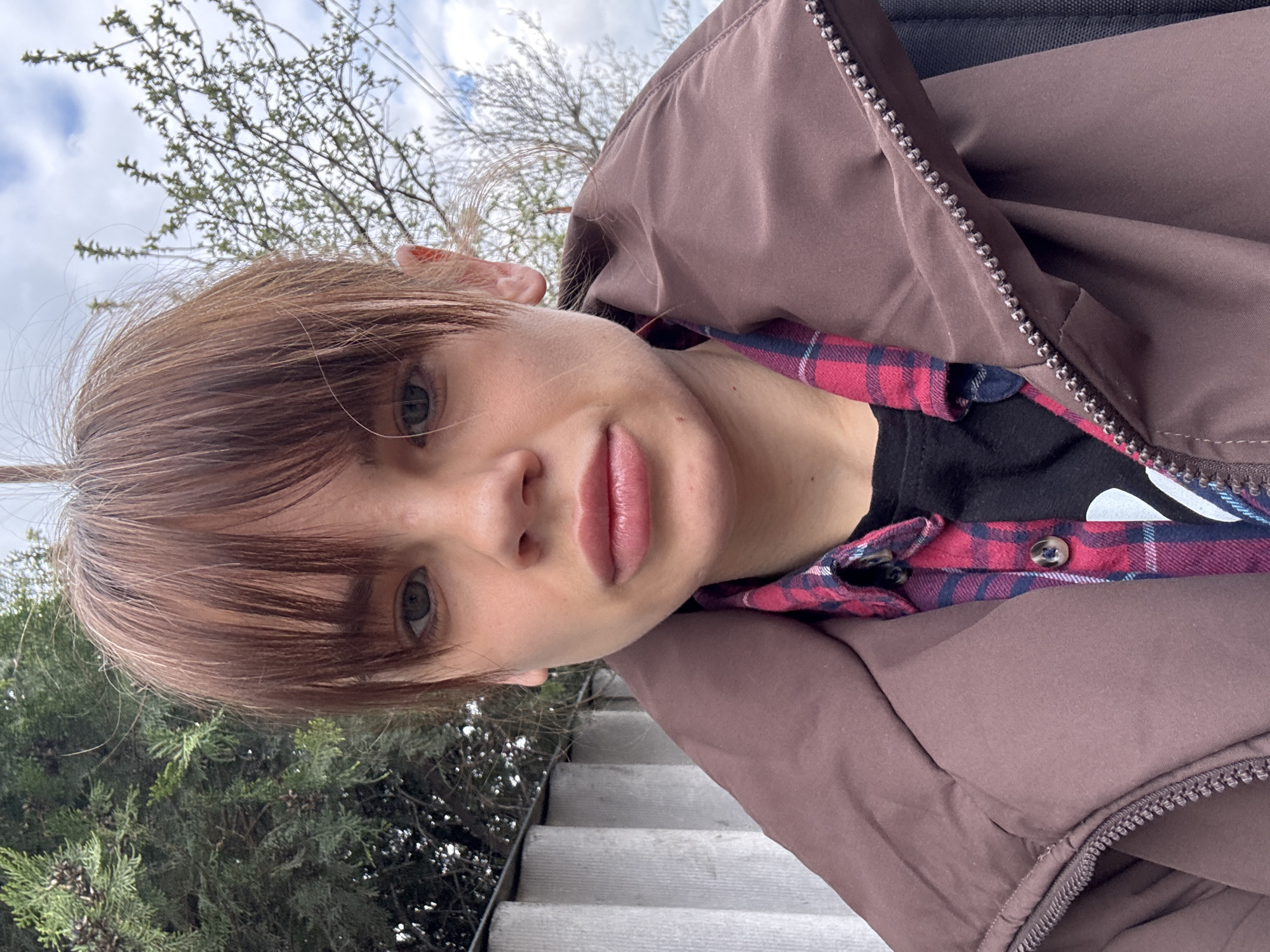 A person is captured in a selfie taken outdoors. The individual has fair skin, light brown hair with bangs, and visible green eyes. The person is wearing a brown puffer jacket over a black shirt and a red and blue plaid shirt. In the background, there are trees and a partly cloudy sky. The person appears to be standing on or near a set of concrete steps. Without additional information or local knowledge, it's impossible to confirm if the photo was taken in Rostov-on-Don. The image provides no visual cues to definitively place it there.