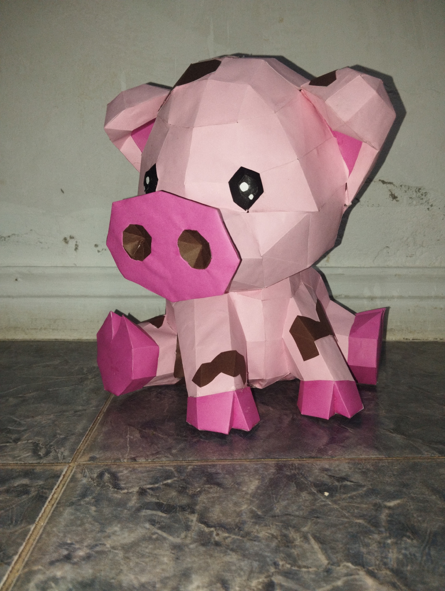 A handcrafted, pink polygonal pig figurine with brown spots is positioned on a gray tiled floor against a light gray wall. The pig is sitting with its legs extended in front, and it has a pink snout with brown nostrils. The figurine's eyes are black with white reflections. The environment indicates an indoor setting. There are no visible people or explicit indications of the city of El Valle del Espíritu Santo, Venezuela.