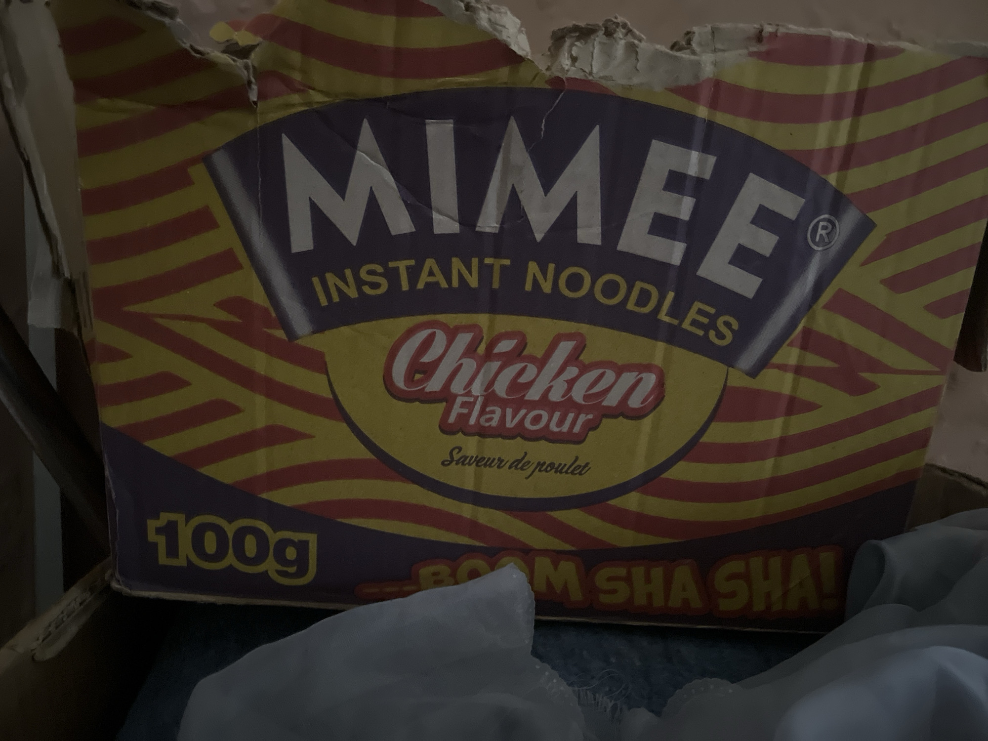 A torn cardboard box with the brand name "Mimee Instant Noodles" is visible. The box is decorated with yellow and red stripes and features a yellow circular logo with "Chicken Flavour" written in red cursive and "Saveur de poulet" in smaller text below. The box also displays "100g" and "BOOM SHA SHA!". The scene appears to be indoors, possibly in Jalingo, Nigeria, with a dark background and a piece of white fabric in the foreground. The lighting is dim, casting shadows on the scene.