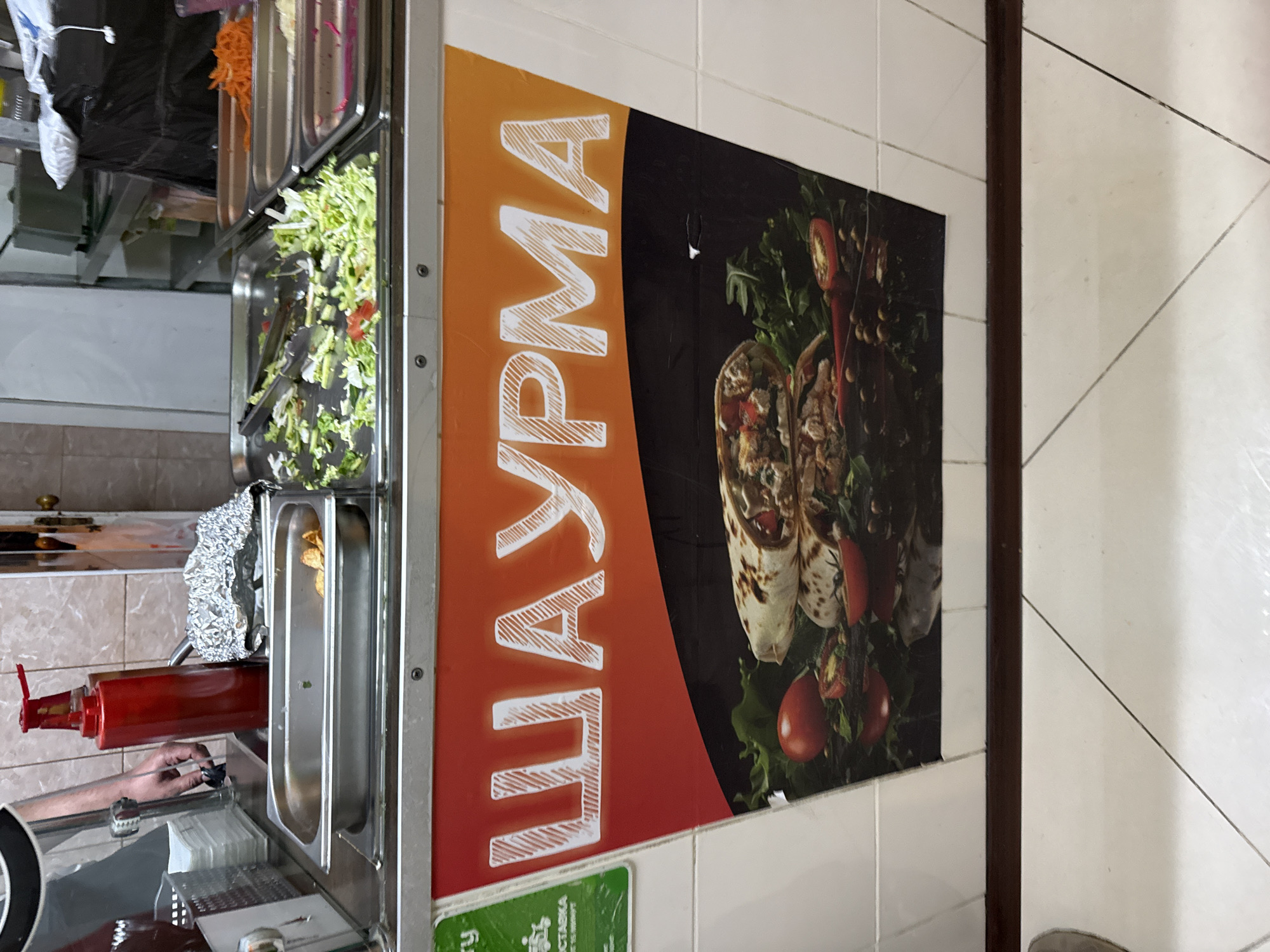 The image depicts a food stall, likely a shawarma stand, in Saint Petersburg, Russia. The foreground features a large, vibrant banner advertising "ШАУРМА" (SHAURMA) with a photographic depiction of shawarma wraps, tomatoes, and greenery. Behind the banner, stainless steel trays are visible, holding various ingredients such as chopped lettuce, tomatoes, shredded carrots, and what appear to be french fries. A red ketchup dispenser stands prominently, and a person's arm and hand, likely that of a vendor, are visible reaching towards it. The background reveals a tiled wall and shelving units, suggesting a commercial food preparation area. The overall impression is that of a busy, functional food service establishment.