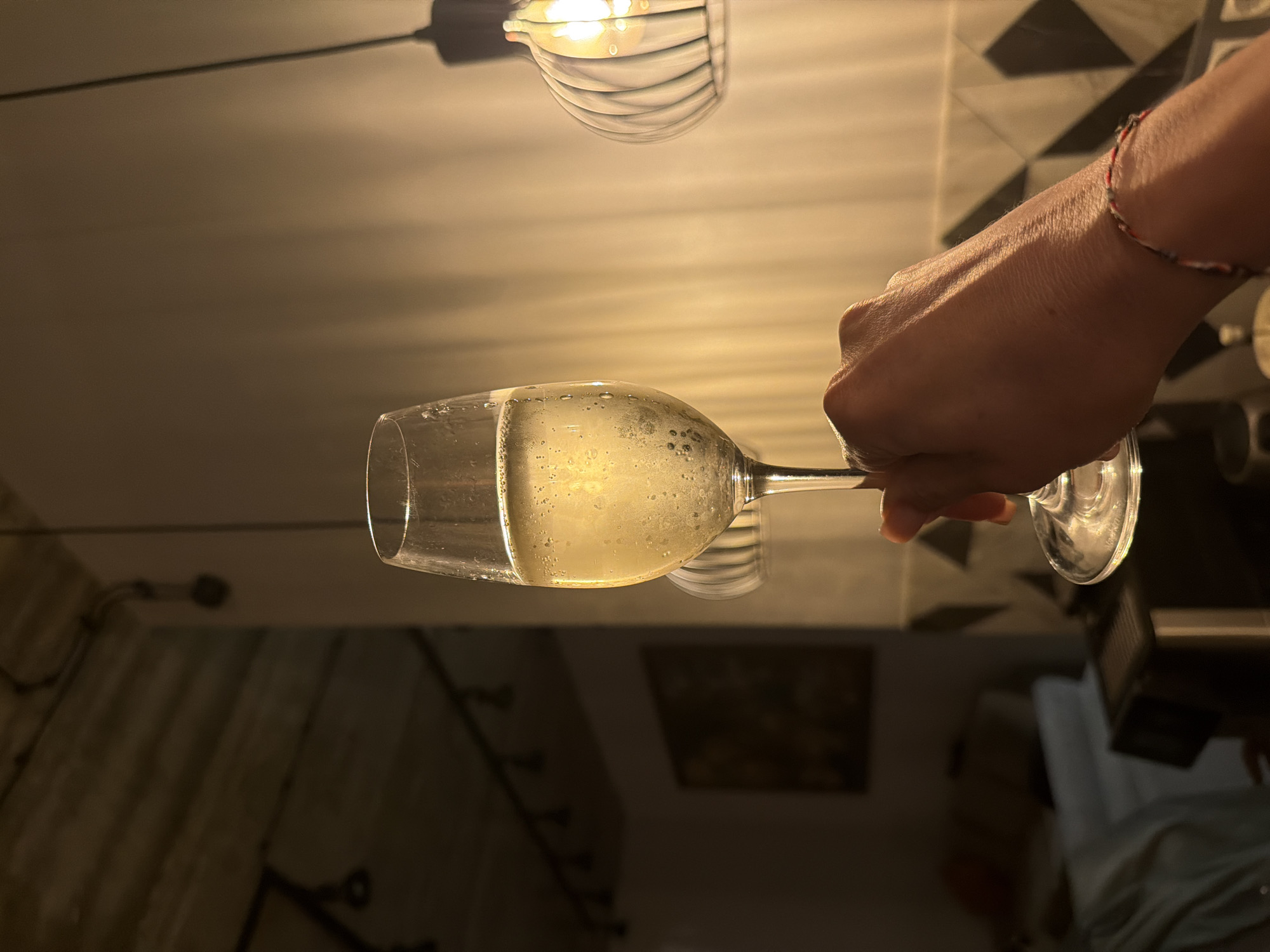 A hand holds a half-full glass of a clear, bubbly liquid, likely champagne or sparkling wine. The glass is illuminated from above by a modern light fixture with a wire cage design. The background shows a mix of elements, including what appears to be a decorative wall pattern, suggesting an indoor setting, possibly a bar or restaurant. There is no clear evidence to confirm that this image was taken in the city of Ryazan, Russia. Therefore, this detail cannot be validated.