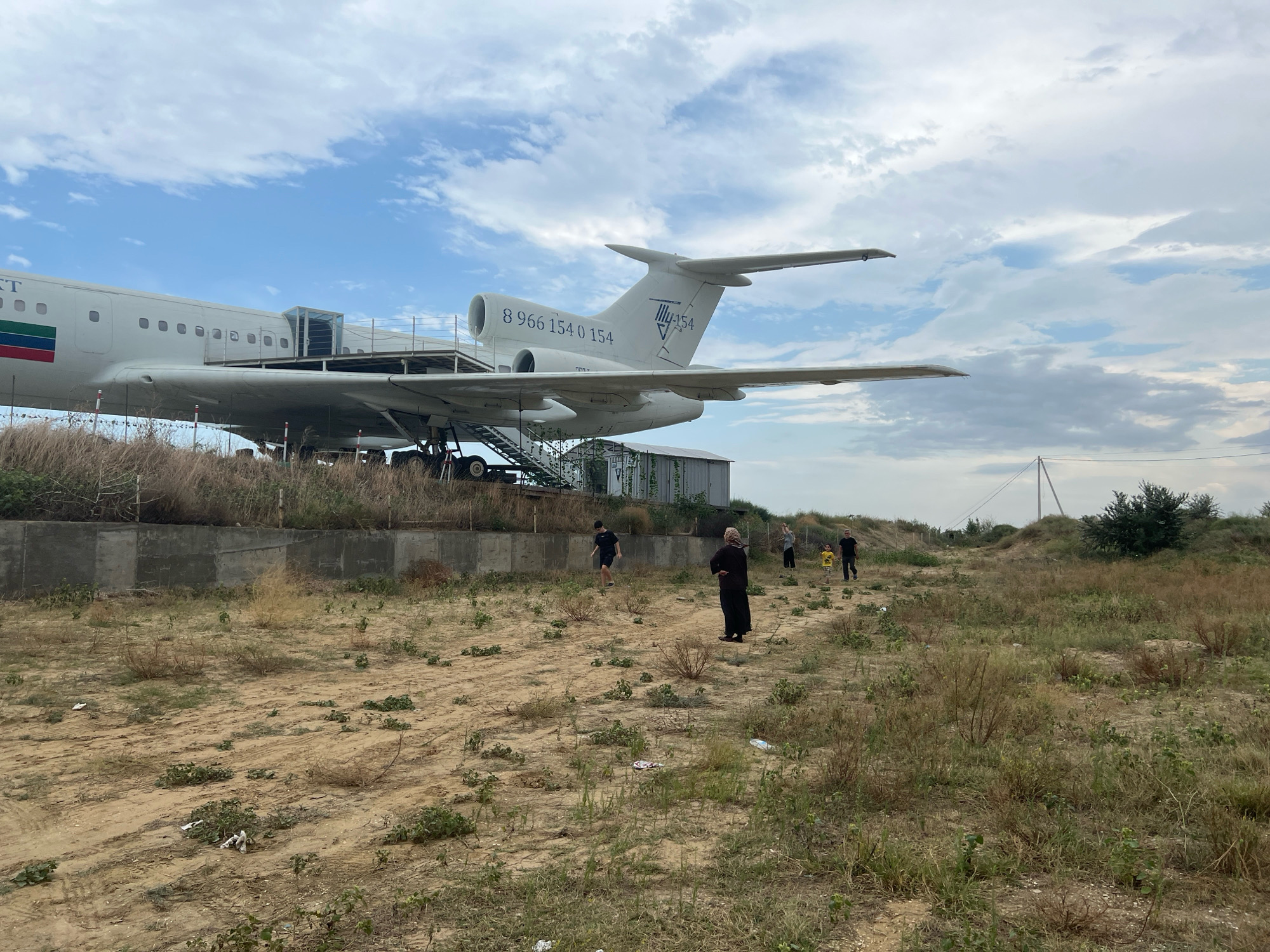 The image shows a white airplane, identified as a Tupolev Tu-154 by the markings "8 966 154 0 154" and "154" on its tail, situated outdoors in a dry, grassy environment. The plane is elevated on supports, with a platform and stairs leading to its fuselage, suggesting it might be repurposed as a structure or exhibit. The location is identified as Manaskent, Russia. Several people are visible in the foreground, walking on the dirt and dry grass. One person in a dark headscarf and long skirt stands facing away from the camera. Another person in a black t-shirt and shorts walks away from the plane. Two other individuals, a person in a light grey t-shirt and another in a yellow shirt, are also present. The background features a cloudy sky, a concrete wall, and sparse vegetation, with a fence and a utility pole visible in the distance.