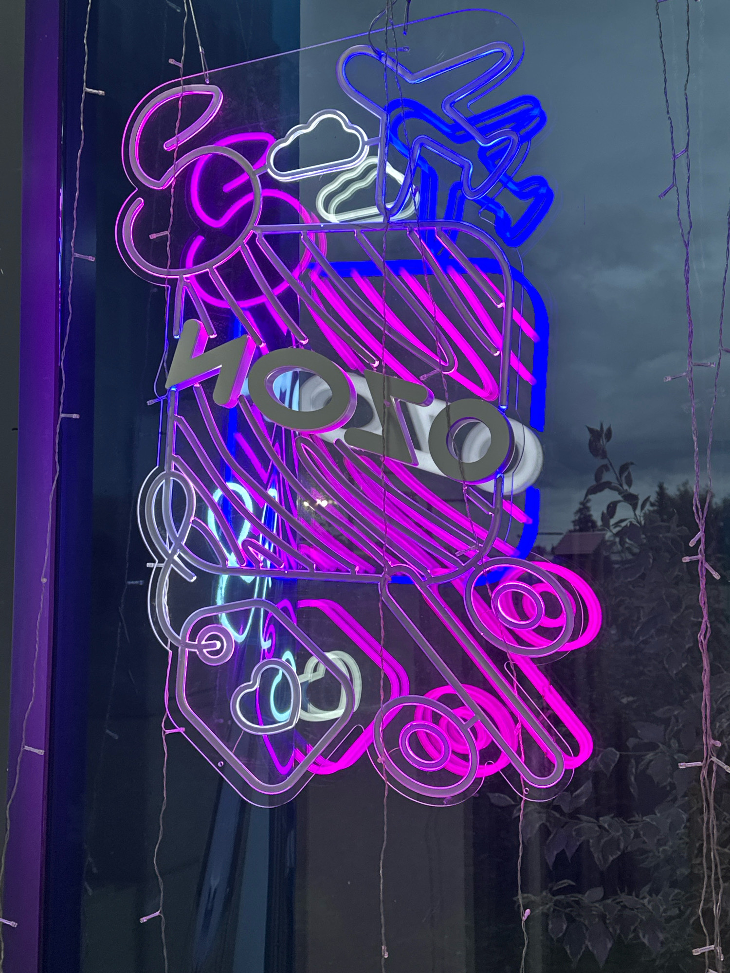 This is a vertical, eye-level, medium shot of a neon sign in a window at night. The sign is a complex design made of pink and blue neon tubes against a dark, reflective background. The sign appears to be comprised of multiple elements, including what looks like a stylized sun with rays, a cloud, an airplane, a gift box with diagonal lines, and various circular and heart shapes. In the center of the sign, in a lighter color, are the letters "MOTO". Small, white fairy lights hang vertically to the left and right of the neon sign. The reflection in the window shows trees and possibly a building, suggesting an outdoor setting. The overall impression is of a vibrant and dynamic display. This image was taken in Vidnoye, Russia.