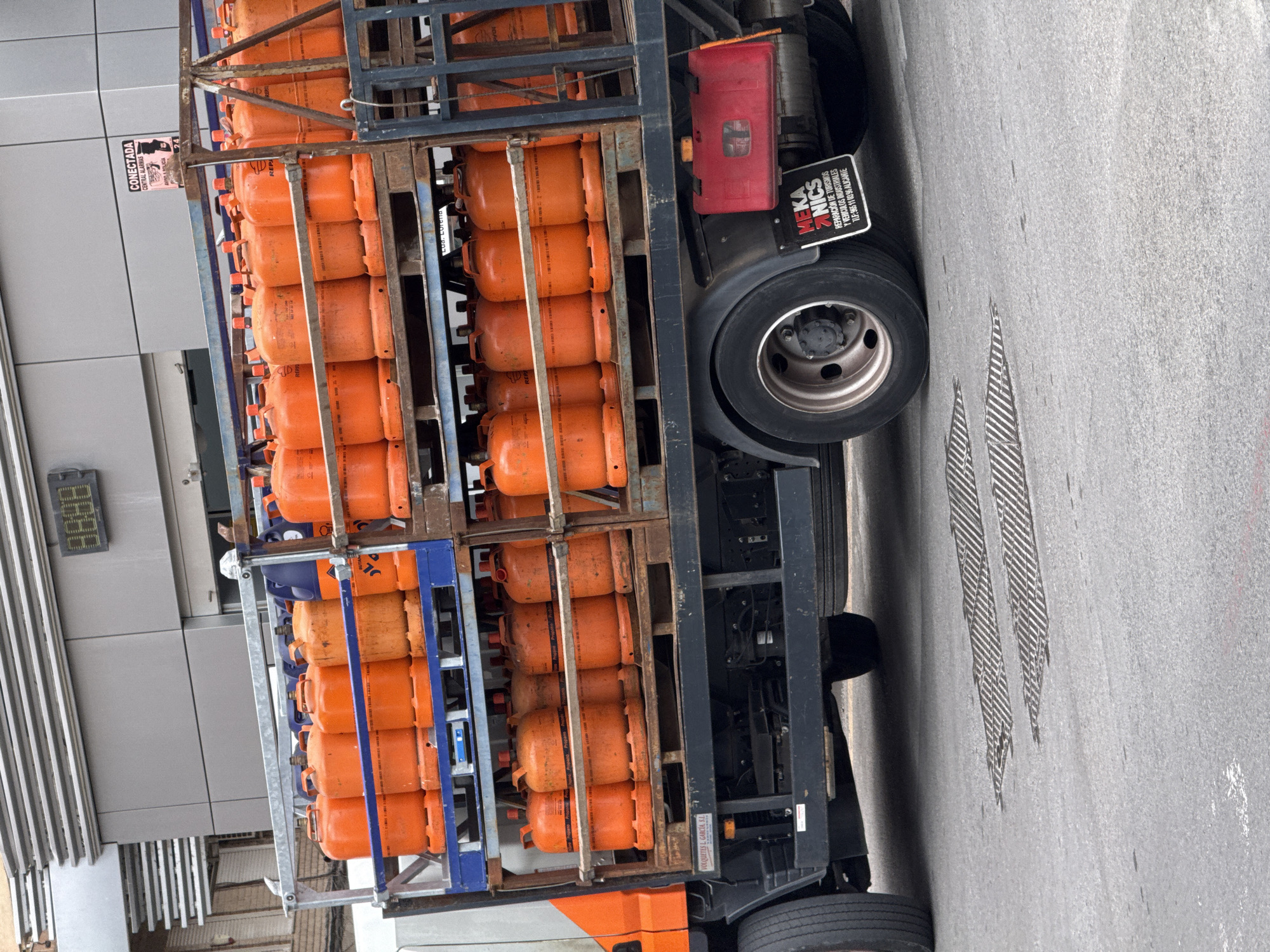 The image shows a truck transporting numerous orange propane tanks in an urban setting. The tanks are stacked in metal frames on the truck bed. The truck has a sign that reads "MEKA ANICS REPARACION DE TURISMOS Y VEHICULOS INDUSTRIALES TLF: 965 11 03 96 ALICANTE" indicating its business as a repair shop. The background includes a gray building with some type of digital display. The location is Unknown, Unknown.