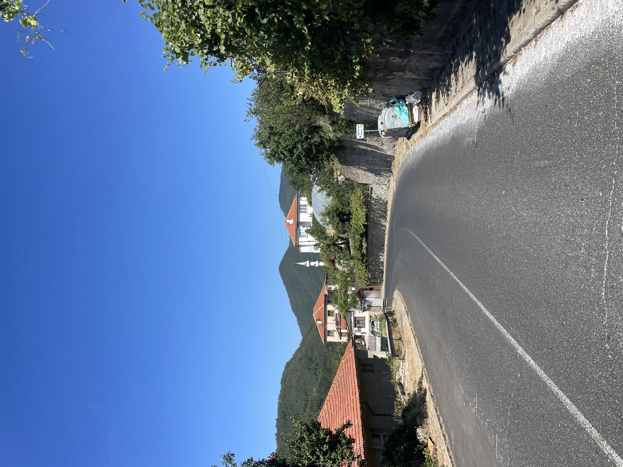 The image depicts a rural road curving through a village under a clear blue sky. On the left, a house with a red tile roof is visible, adjacent to a grey, low-lying structure. The road continues to curve right, leading towards a cluster of buildings further in the distance. Among these buildings, a prominent white structure with a red-tiled roof and a minaret can be seen, suggesting a mosque. The background is dominated by lush green, tree-covered hills.
To the right of the road, a concrete wall lines the side, with a trash receptacle overflowing with debris, including a blue tarp, positioned next to it. A signpost with Cyrillic lettering and an arrow pointing left is mounted on the wall. The area is characterized by natural vegetation and a sense of a peaceful, somewhat aged, village environment. The image seems to be taken in Oryahovets, Bulgaria, given the visual elements present. There are no people visible in the image.