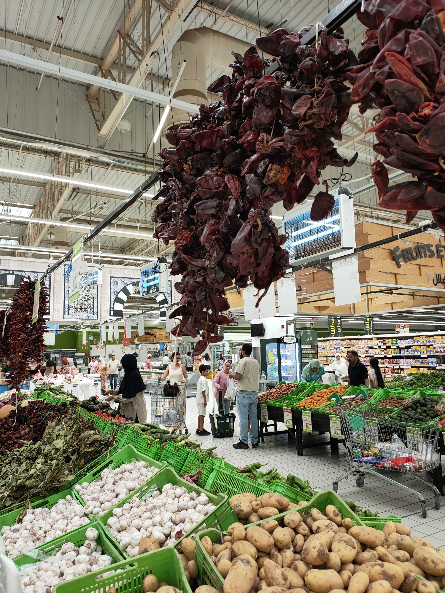 The image depicts the interior of a supermarket in Cebalat, Tunisia. In the foreground, several crates overflow with fresh produce, including large quantities of garlic bulbs and potatoes. Beyond these, a variety of fruits and vegetables are displayed in green crates, such as apricots, peaches, kiwis, and cucumbers. Hanging above the produce sections are clusters of dried red peppers, adding a rustic element to the market.
In the middle ground, shoppers navigate the aisles. A woman wearing a dark headscarf is seen interacting with produce, while other individuals, including a man in a black polo shirt and a young boy, are also present. Shopping carts are visible, indicating ongoing purchases.
The background reveals the typical supermarket environment with shelves stocked with packaged goods, overhead lighting, and various signs, some featuring Arabic script. The overall scene suggests a bustling marketplace where fresh produce is a central offering.