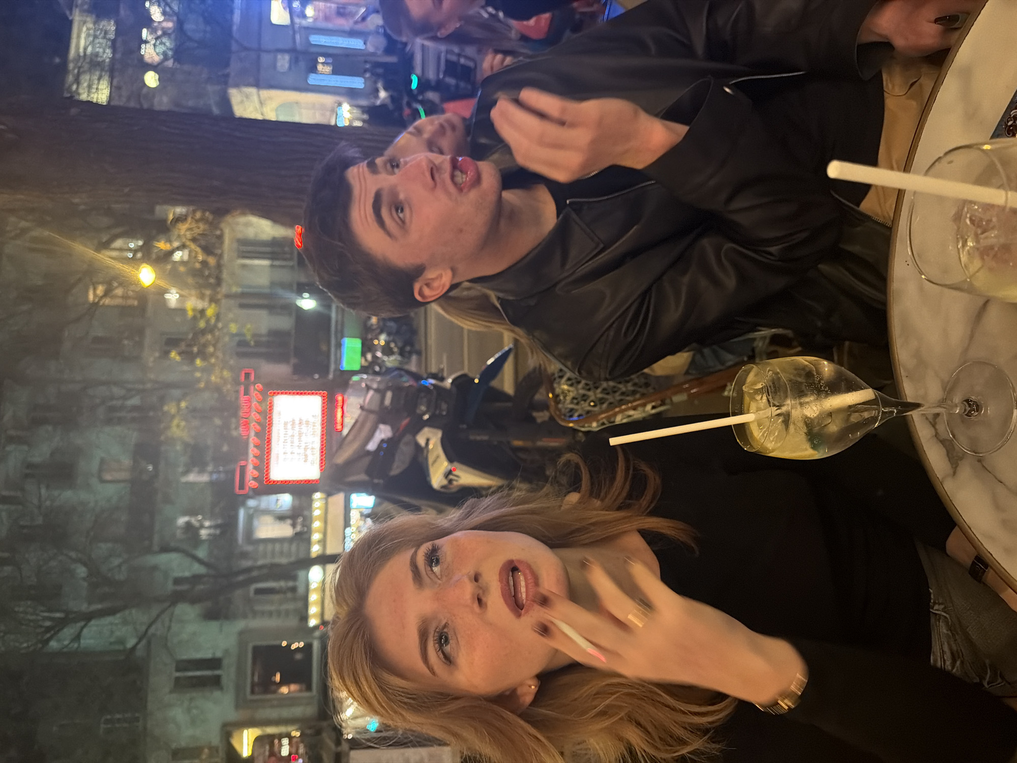 Two young adults, a male and a female, are seated at an outdoor café table. The female is holding a cigarette near her mouth. Both have drinks with straws. The male is wearing a black leather jacket. The setting appears to be Paris, France, indicated by the architecture and neon signs visible in the background.