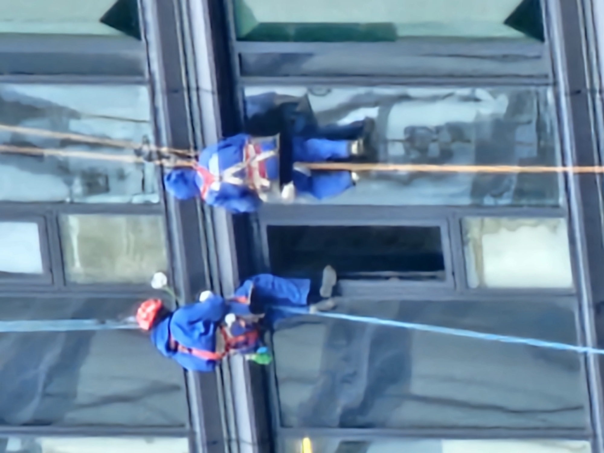 Two individuals, dressed in blue work suits and safety harnesses, are suspended from ropes attached to a building, likely engaged in window cleaning or exterior maintenance. The person on the left wears a red hard hat. They are positioned against a modern building with large windows, some of which appear to have internal blinds or curtains. The scene suggests an urban environment, but the specific location in Moscow, Russia, cannot be determined from the visual information alone.