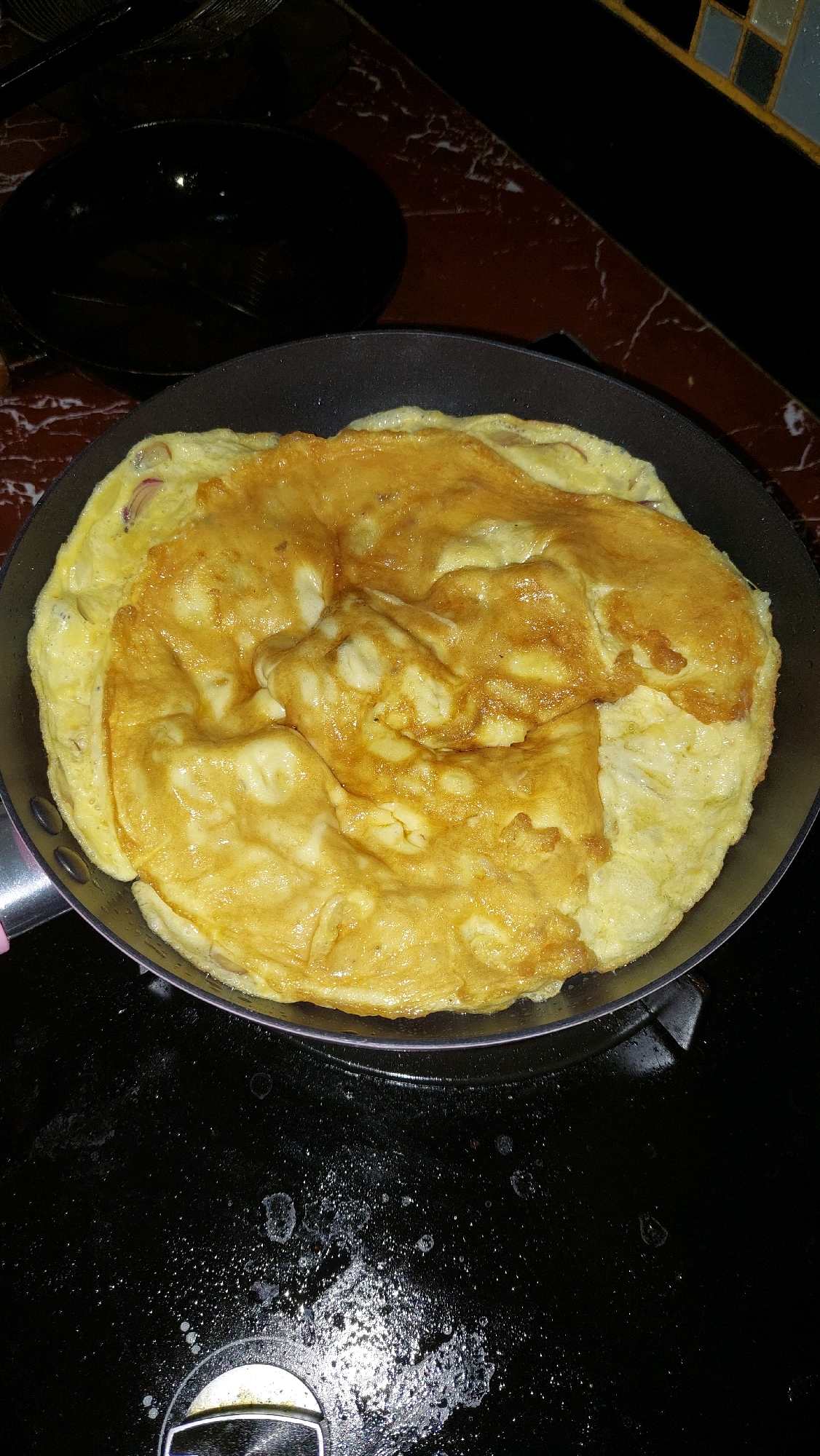 A close-up, overhead shot captures an omelet being cooked in a black frying pan on a black stovetop. The omelet is golden brown and appears folded in half, with some slightly browned edges. The pan has a pink handle and is set on a stovetop that has visible water droplets and stains. In the background, a blurred dark surface, possibly a countertop with a marble pattern, is visible, along with part of a black wok or pot. The scene suggests a home kitchen setting in Bekasi, Indonesia, where food preparation is taking place.