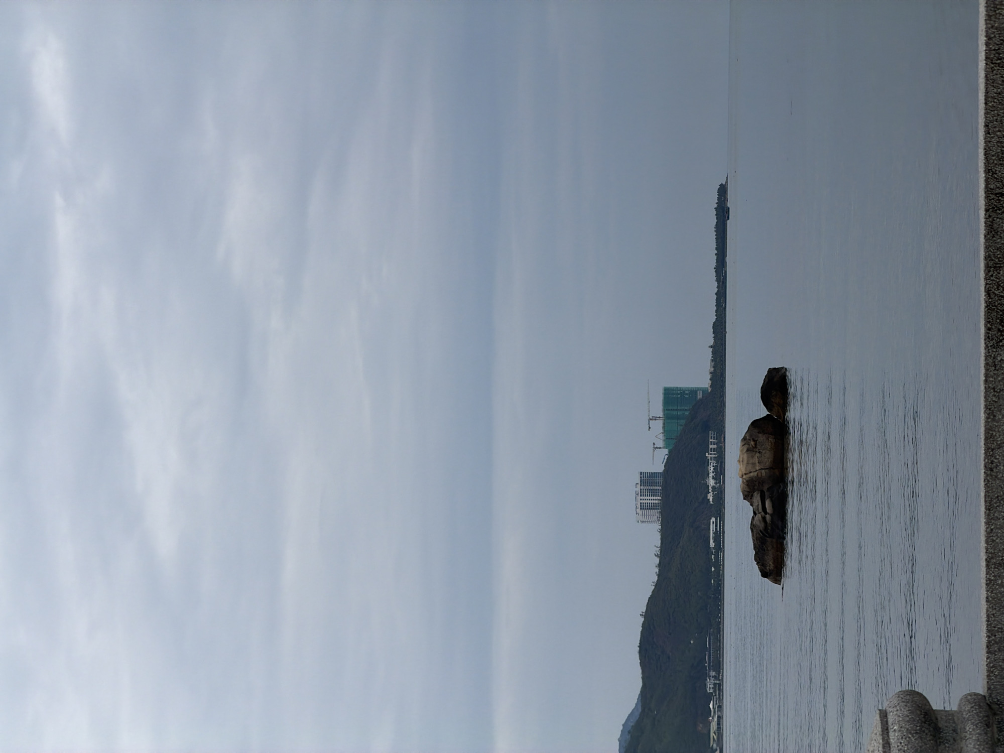 The image shows a landscape in Bac Nha Trang Ward, Vietnam, featuring a large rock formation in the water with a mountain and buildings in the background. The sky is overcast, and the water is calm. A portion of a stone barrier or wall is visible in the foreground on the right.