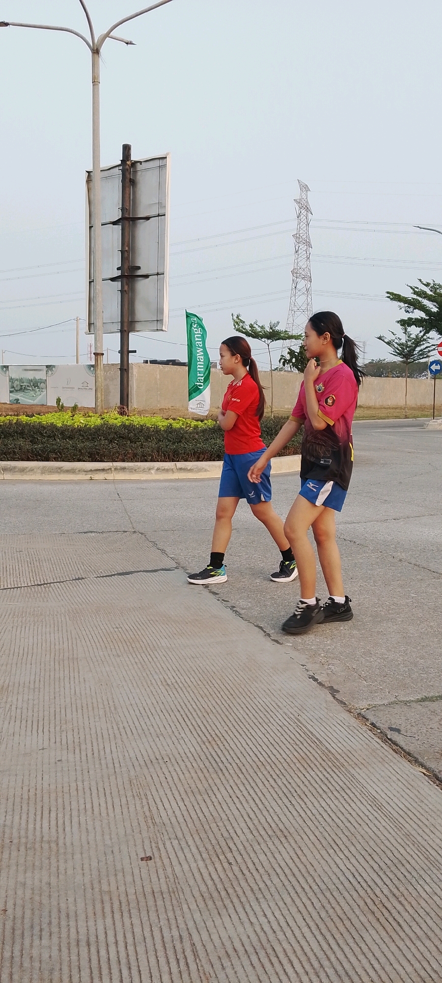 Two young females are walking side-by-side on a paved road with concrete grooving in Satriamekar, Indonesia. The female on the left is wearing a red short-sleeved shirt with blue shorts and black athletic shoes with white accents. She has her arms crossed in front of her. The female on the right is wearing a pink and black short-sleeved shirt with blue shorts and black athletic shoes. She is looking to her right and touching her chin with her right hand. Behind them, to the left, is a tall metal lamppost and a large rectangular sign mounted on a pole. To the right of the sign, a green flag with white text is visible, partially obscured by the females. Further in the background, there is a large metal tower with power lines, trees, and a light blue sky. The ground in front of them is a paved road, and on the left side of the road, there is a median with green shrubs and some construction signage. The lighting suggests it is daytime, possibly late afternoon.