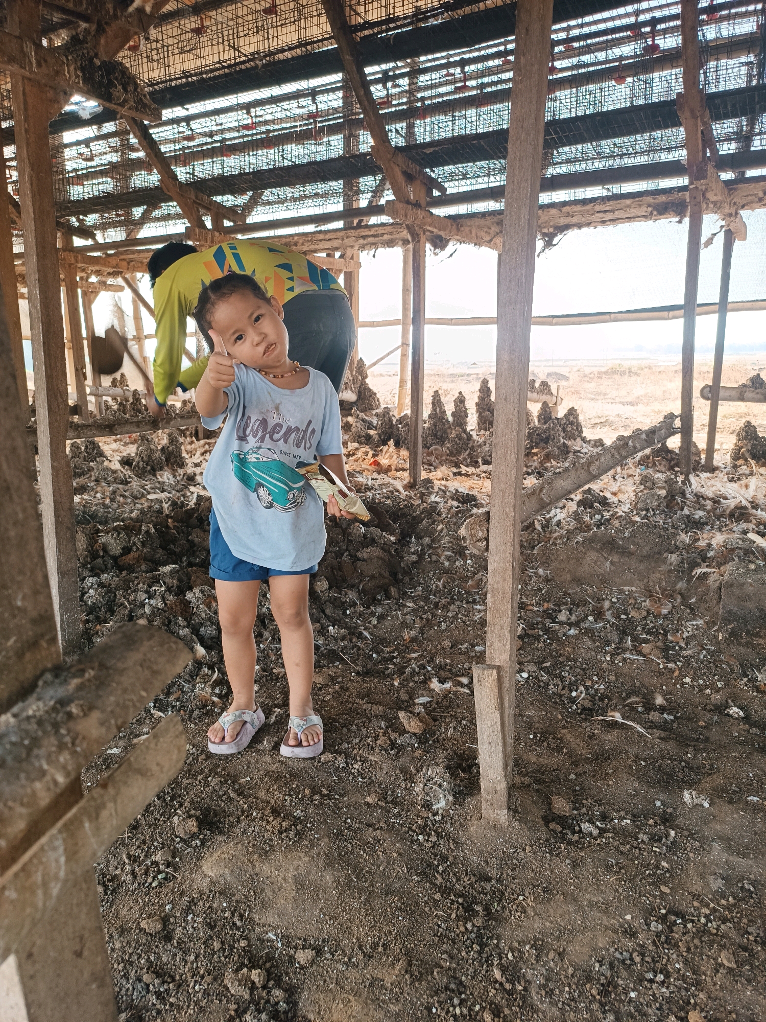 A young child, wearing a light blue t-shirt and blue shorts, stands in the foreground of a building in Tambakromo, Indonesia. The child is giving a thumbs-up gesture and holding a small item in their right hand. Behind the child, another person, wearing a lime green long-sleeved shirt, is engaged in some activity. The building appears to be an open-air structure with wooden beams and a metal roof. The ground is covered in dirt and debris. The background suggests an outdoor environment with dry vegetation.