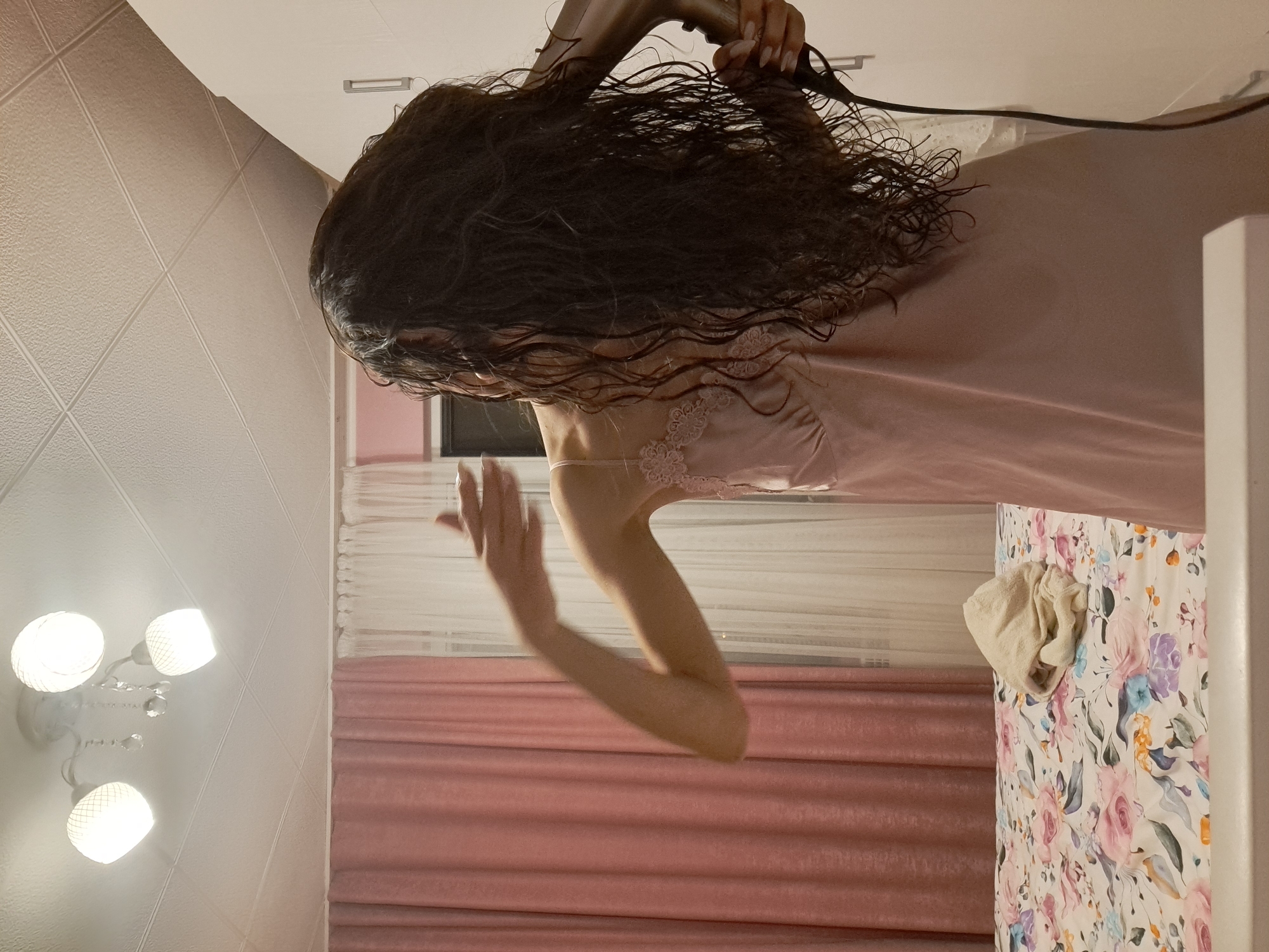 A young woman with long, dark, wet hair is drying her hair with a hairdryer in a bedroom. She is wearing a pink slip dress with lace trim. Her right arm is raised, and her left hand is holding the hairdryer. The room is decorated with pink and white curtains, a floral-patterned bedspread, and a white ceiling with three lights. A white towel is placed on the bed. The scene appears to be within a residential property located in Negovan, Bulgaria.