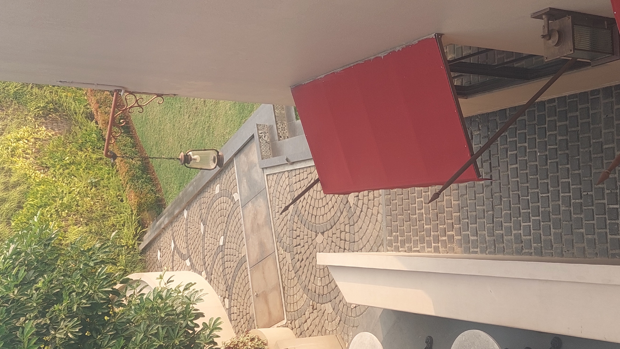 This image depicts an outdoor area with a focus on architectural elements and landscaping. In the foreground, a red awning extends from a beige-colored building. Below the awning, a dark brick-paved walkway is visible. To the left, a white structure with a curved edge is present, and further left, a round table can be seen.
The midground features a pathway paved with rounded cobblestones arranged in a circular pattern. Adjacent to this pathway is a well-maintained lawn and a dense row of manicured bushes. A hanging lantern with a glass shade is suspended from an ornate metal bracket attached to the building.
The background shows lush green foliage with a sunlit effect, suggesting daylight. The overall scene appears to be an exterior walkway or patio area, possibly part of a restaurant, hotel, or residential complex, located in an urban environment like Cileungsi, Indonesia. There are no people visible in the image.