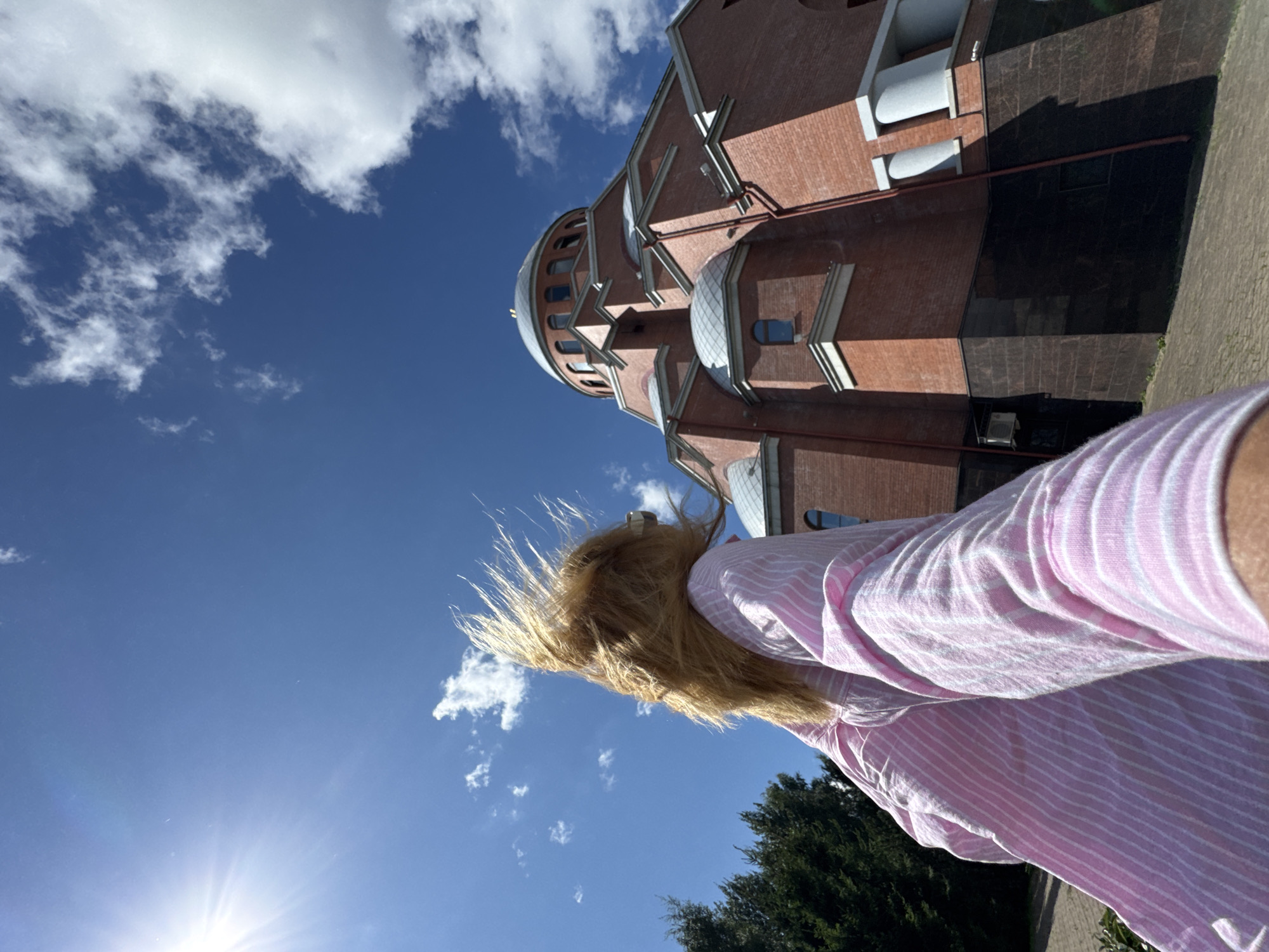 A low-angle, outdoor, wide shot captures a woman with blonde hair, her face obscured by wind-blown hair and the camera angle, standing in front of a large, red brick building with a dome. She wears a pink and white striped shirt. The sun shines brightly from the upper left corner, casting a lens flare. The sky is a clear blue with scattered white clouds. A large evergreen tree is visible on the left side of the frame. The building appears to be a religious structure, possibly a church or cathedral, with arched windows and decorative architectural elements. The ground in front of the building is paved with cobblestones. The image appears to have been taken on a sunny day. The architectural style suggests it may be located in Saint Petersburg, Russia.