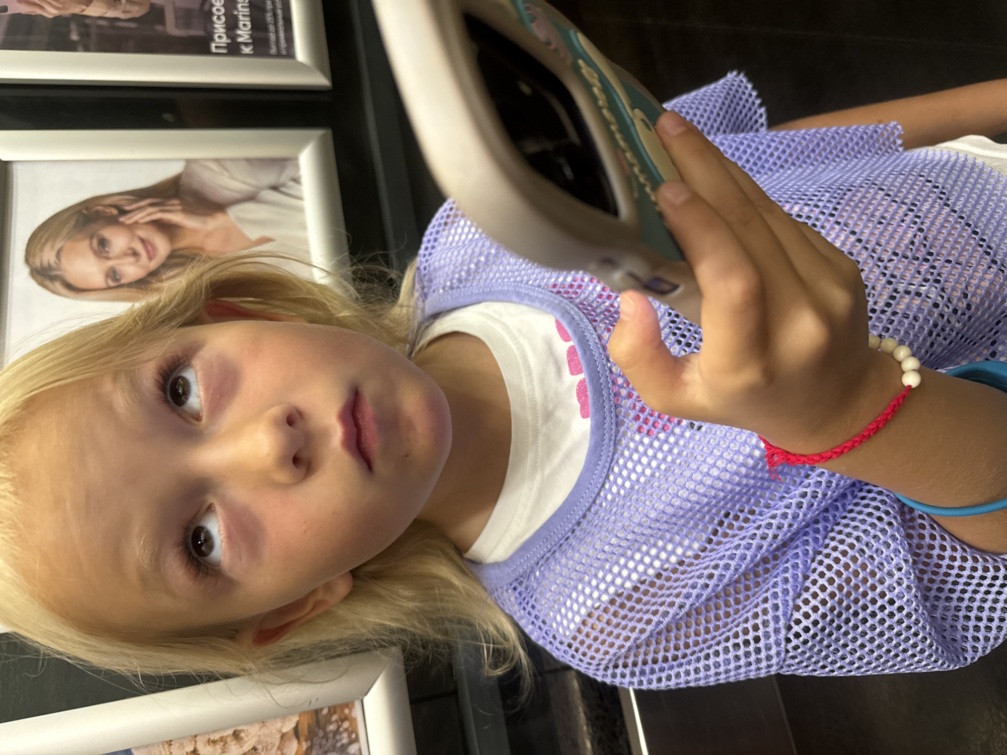 A young blonde girl with blue eyes looks upwards, her phone in her right hand. She is wearing a white t-shirt with a pink graphic and a sheer lavender mesh top. She has a red bracelet with white beads on her wrist. Behind her, a poster of a woman with blonde hair is visible. The scene appears to be indoors, possibly in a public place in Yalta, Ukraine.