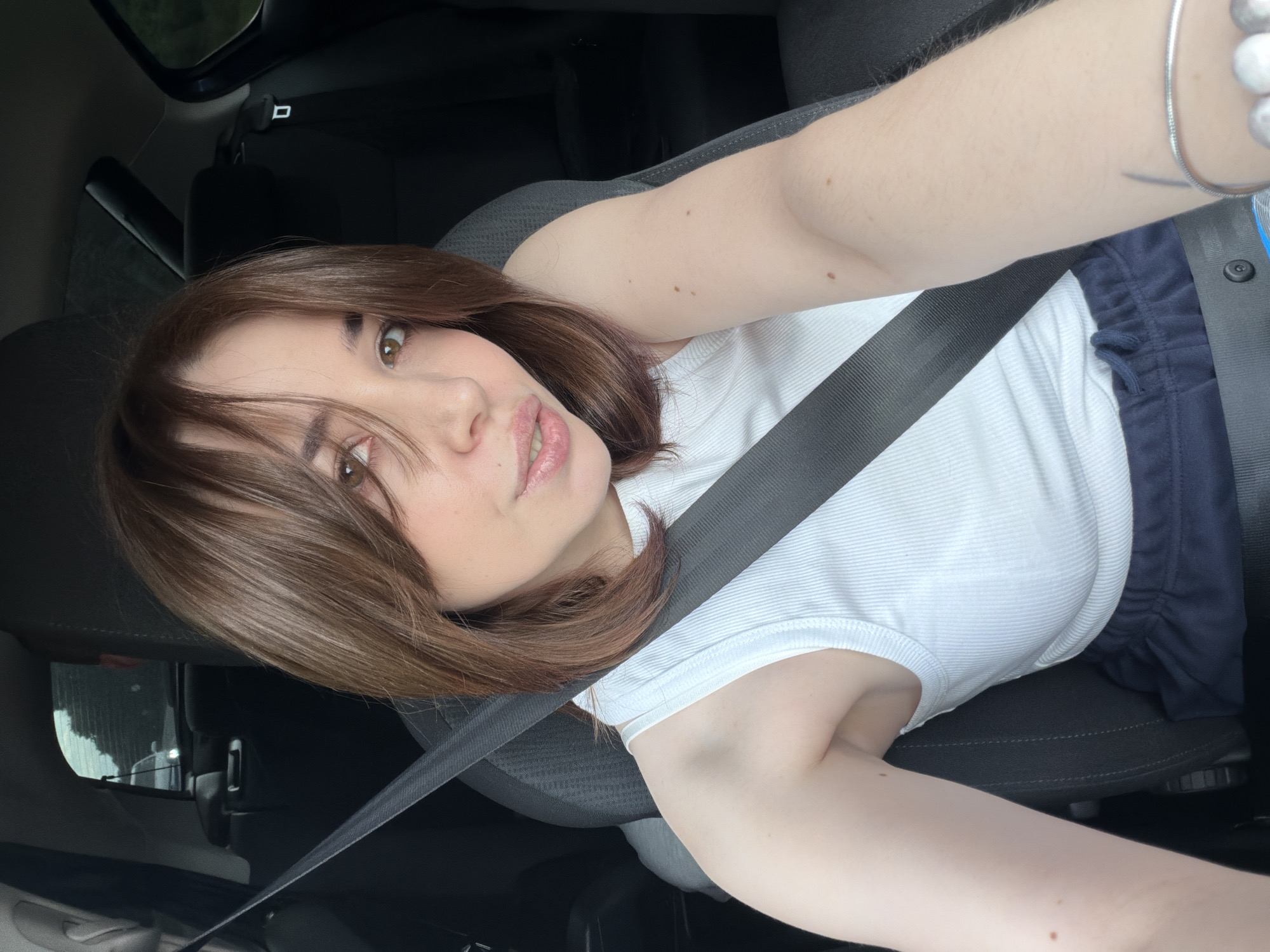 This is a portrait-oriented, medium shot image of a young woman with shoulder-length brown hair, taken from a slightly high angle. She is seated in the driver's seat of a car, wearing a white ribbed tank top and dark blue sweatpants. A black seatbelt crosses diagonally over her chest, and her left arm is extended towards the camera. Her right arm is not fully visible.
The woman is looking directly at the camera with a relaxed expression, her lips slightly parted. Her makeup includes foundation, blush, and lip gloss. A thin, silver bracelet is visible on her left wrist, and she has a small tattoo on her inner left forearm.
The interior of the car is visible, with dark gray seats and a black seatbelt buckle. The background is mostly dark, with some light reflecting off the driver's side window, suggesting it might be daytime or that the car's interior lights are on. The overall mood is casual and candid. No text is visible in the image.