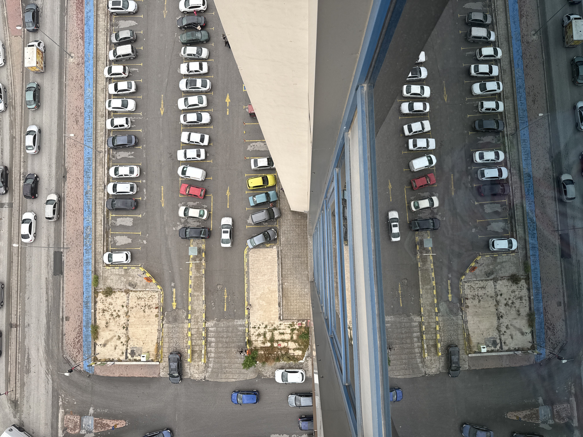 An aerial view captures a parking lot with various cars, a building, and surrounding urban infrastructure in the city of Konya, Turkey. The scene is dominated by parked vehicles, including sedans and SUVs, in different colors and models. Some cars are neatly parked in designated spots, while others are angled, indicating recent or ongoing movement. The parking lot is paved with asphalt and marked with yellow lines for parking spaces and directional arrows. A tall building with reflective windows is partially visible, casting a reflection of the sky and the scene below. In the background, there are glimpses of streets with more cars and pedestrian walkways. The overall environment appears to be a typical urban setting, likely a commercial or residential area.