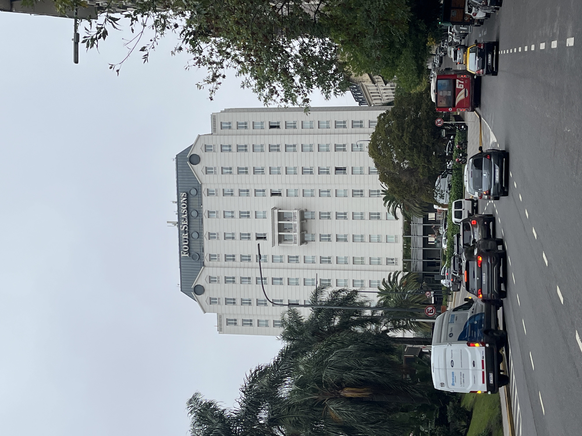 The image shows the Four Seasons Hotel in Buenos Aires, Argentina, from a street view. The hotel is a tall, white building with many windows and a grey roof featuring the "FOUR SEASONS" logo. Palm trees and other greenery are present in front of the building. A street runs alongside the hotel, with various vehicles including cars, vans, and a bus. The traffic appears to be congested. The sky is overcast.