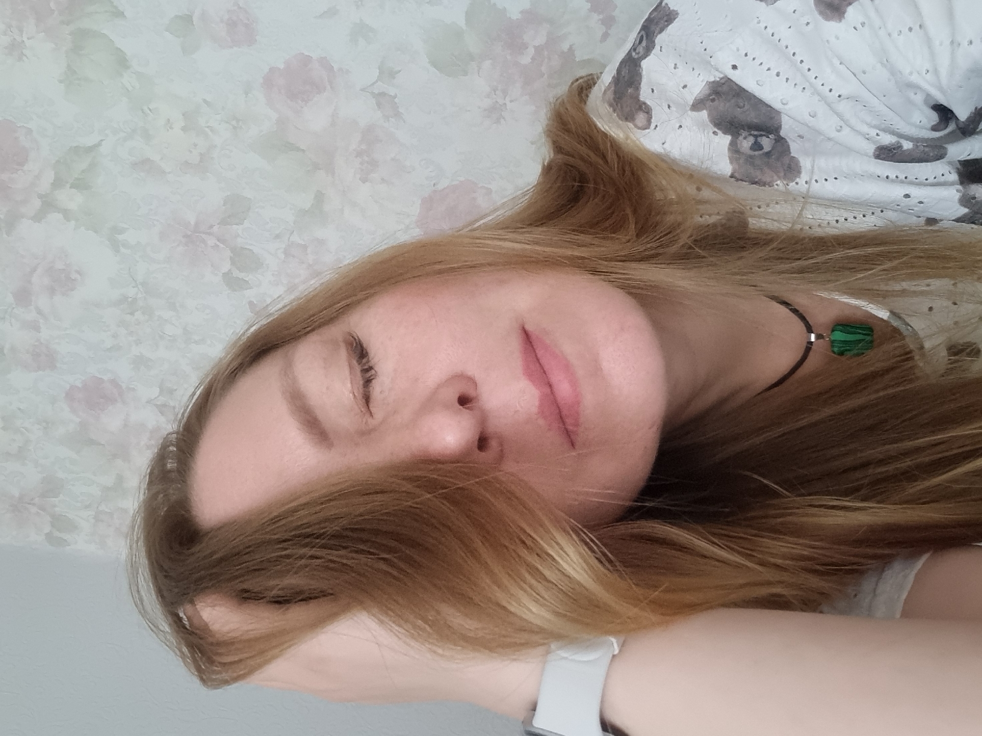 A light-skinned woman with long, blonde hair is depicted in a close-up, portrait-style photograph. She is lying down with her eyes closed and her hair partially covering her face. She is wearing a white smartwatch on her left wrist. She is also wearing a necklace with a green stone. Her head is against a surface that appears to have a floral pattern. The upper right corner of the image shows a white fabric, likely a shirt or blanket, with a pattern of teddy bears on it.
There is no information in the image to determine the location as the city of Chaikovsky, Russia.