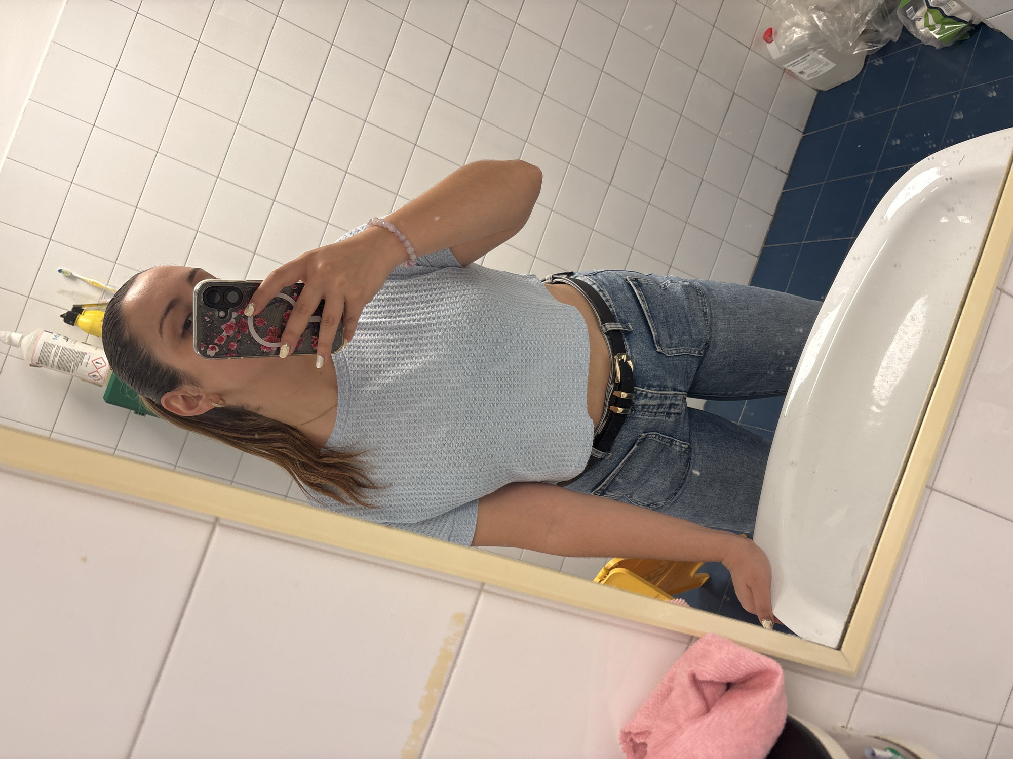 A female individual is taking a selfie in a bathroom setting. The bathroom has tiled walls and a tiled floor with a combination of white and blue tiles. The individual is wearing a light blue waffle-knit top, blue jeans with a black belt, and a beaded bracelet. The individual is holding a cell phone with a floral-patterned case. In the background, there are various cleaning products, including bottles and containers, near the sink. Additionally, a pink towel is visible near the bottom of the mirror. The presence of specific cleaning products and bathroom fixtures suggests this is a residential bathroom. I cannot confirm the location is Barcelona, Spain, from the provided image.