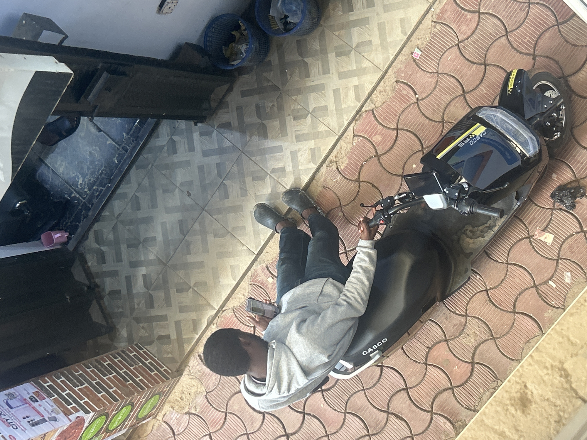 A man sits on a parked black electric scooter in Jos, Nigeria, looking at a phone. The scooter, branded "CASCO," is positioned on red patterned pavement. The man wears black jeans, grey shoes, and a grey hooded sweatshirt. Nearby are areas of grey tiled flooring, bins, and a building with a brick-patterned base.